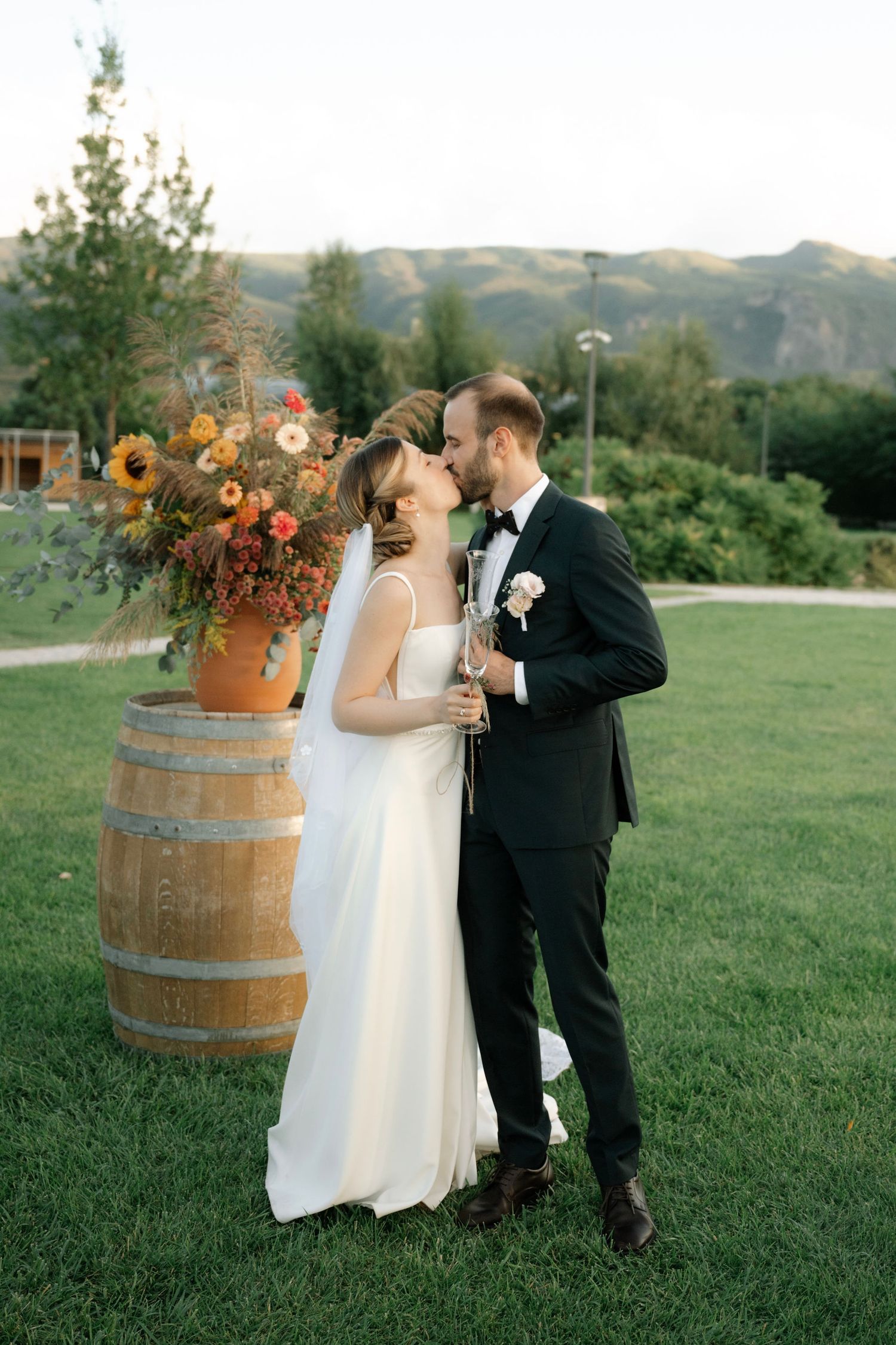 A rustic outdoor wedding scene with wine barrel decor and fall-colored floral arrangements in a mountainous setting.