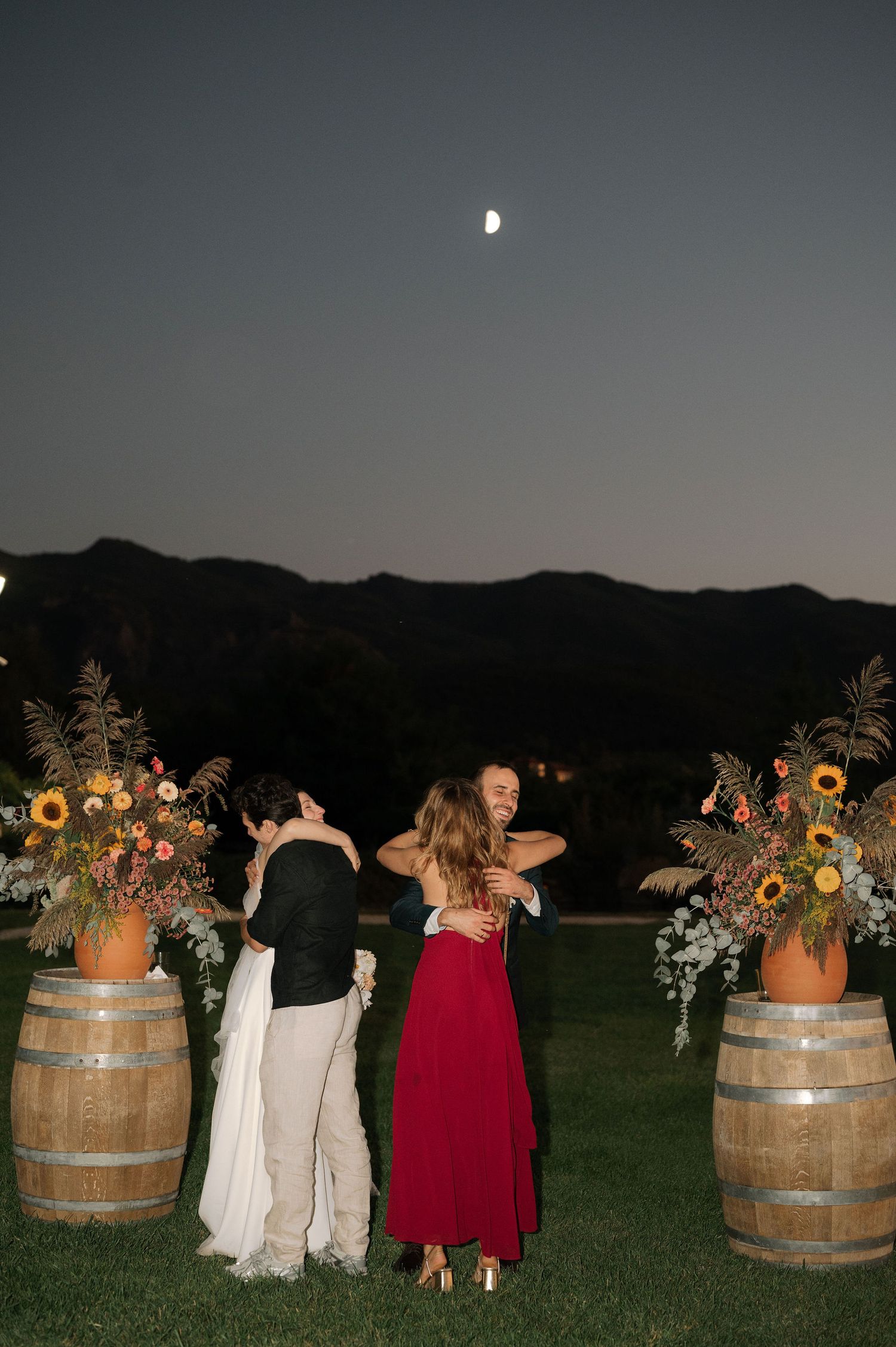 Evening outdoor event with wine barrels and floral arrangements under a moonlit sky in a mountain setting.