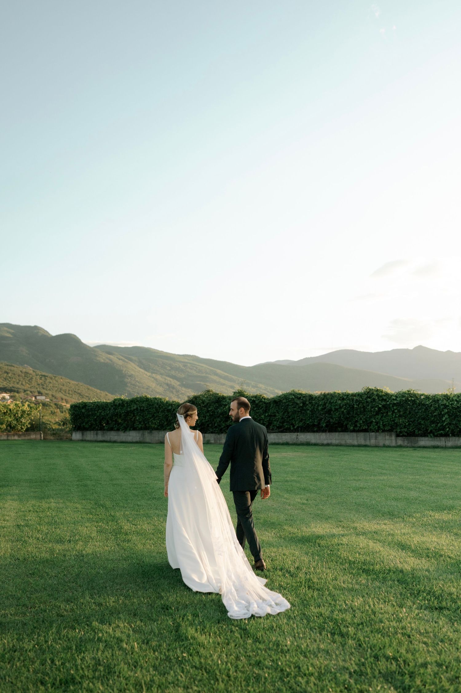 A couple in wedding attire walk hand in hand across a lush green lawn with mountains in the background at sunset.