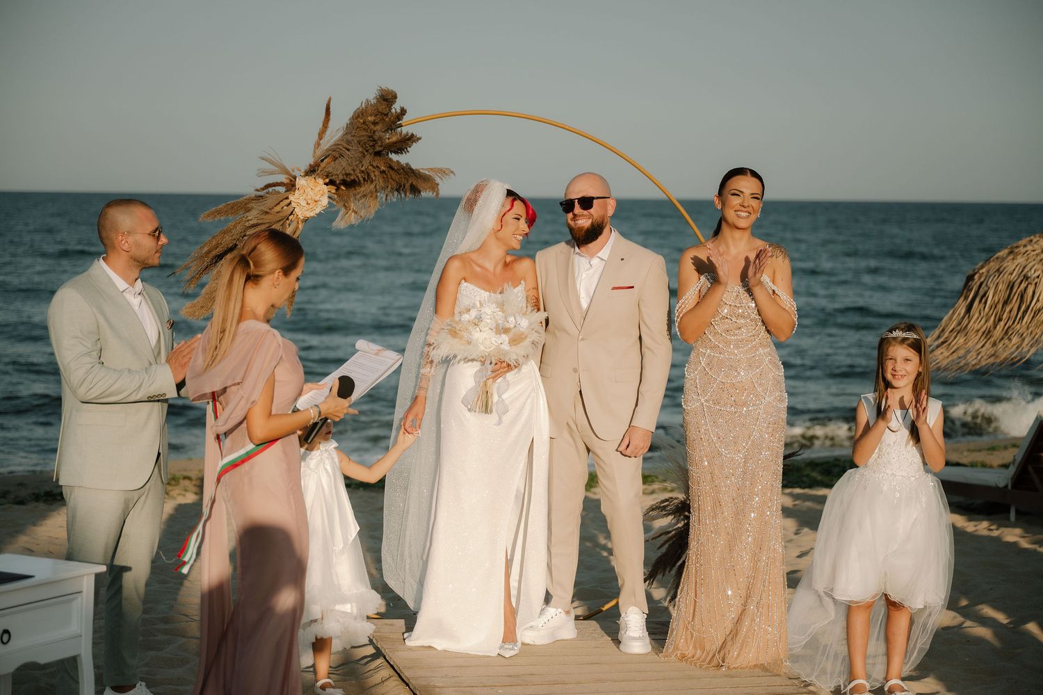 Wedding party in white and beige attire poses together on a sandy beach at sunset with ocean waves in background.