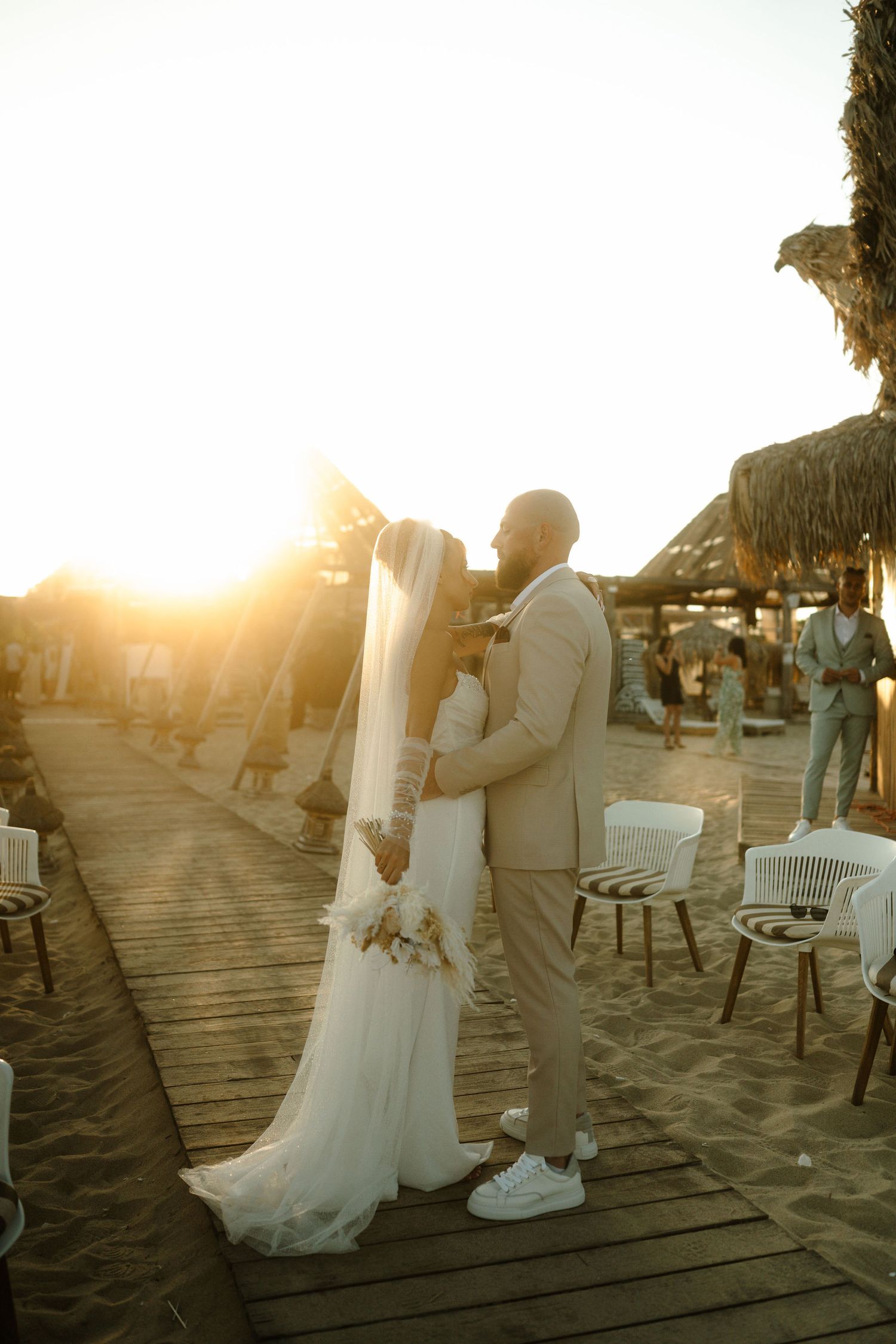 Couple shares a romantic moment on a beachside deck at sunset with palm trees and chairs in the background.