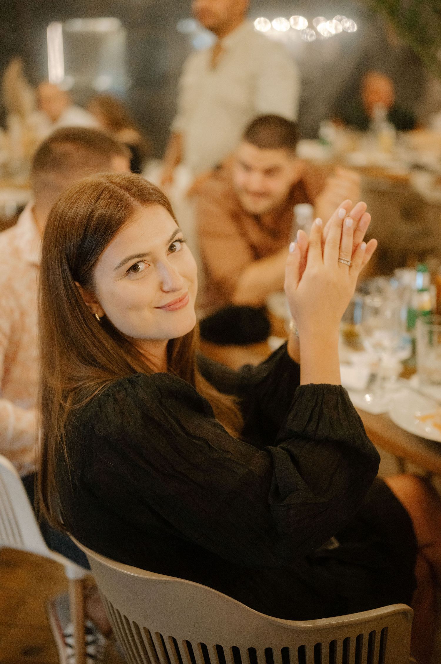 Someone in a black long-sleeve top sits at a dinner table clapping their hands during what appears to be a celebration.