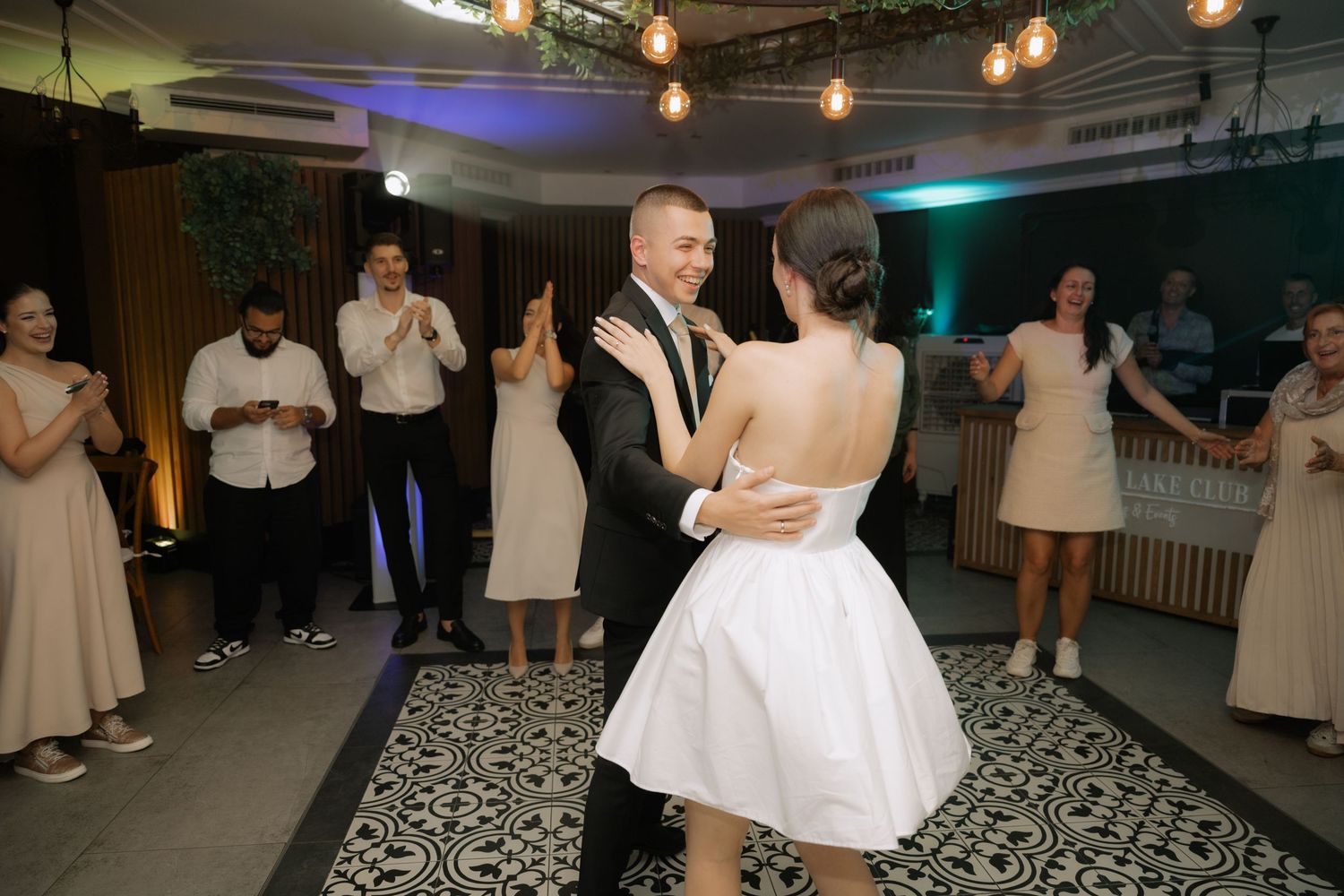 Wedding guests dance on a patterned black and white floor under warm lighting at an indoor reception venue.