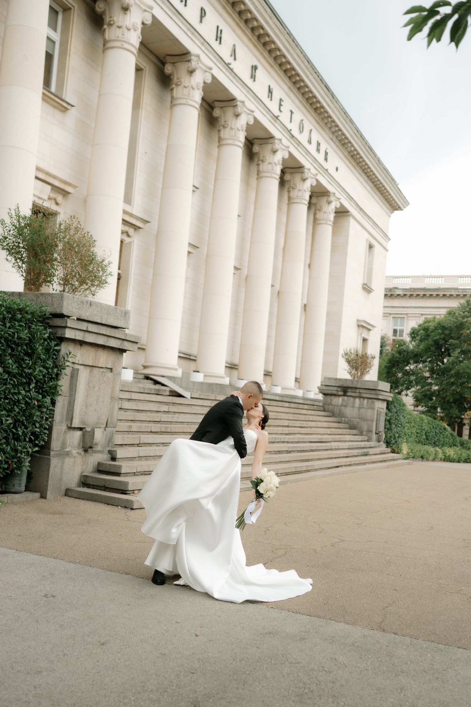A couple shares a romantic moment on the steps of a grand neoclassical building with white columns.