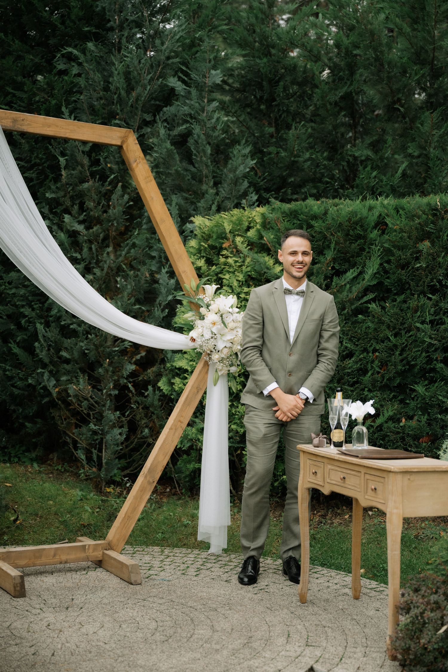 A groom stands under a wooden arch decorated with white fabric and flowers next to a rustic table in an outdoor garden setting.