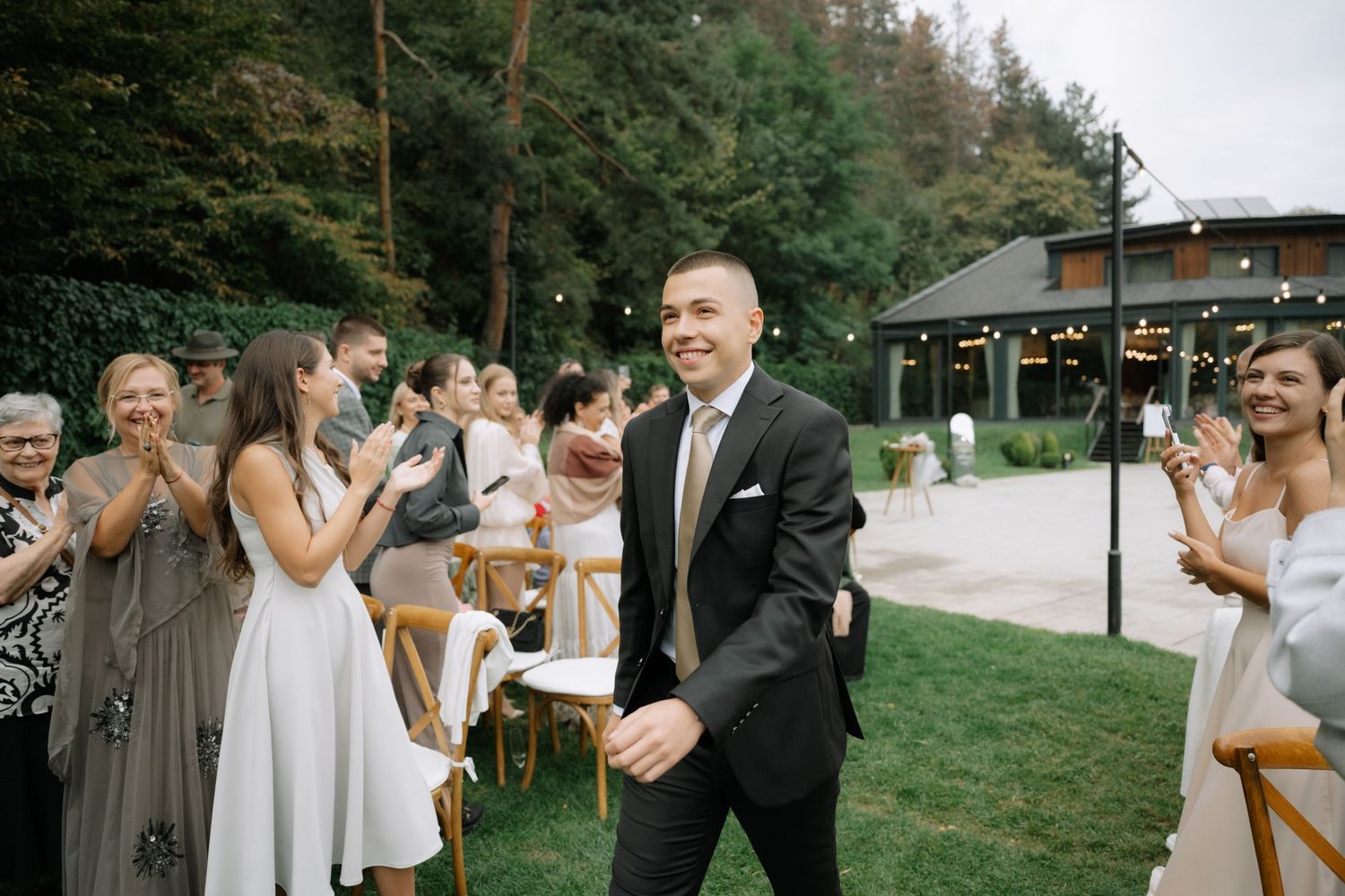 Outdoor wedding celebration on a grassy lawn with guests in formal attire applauding during the ceremony.