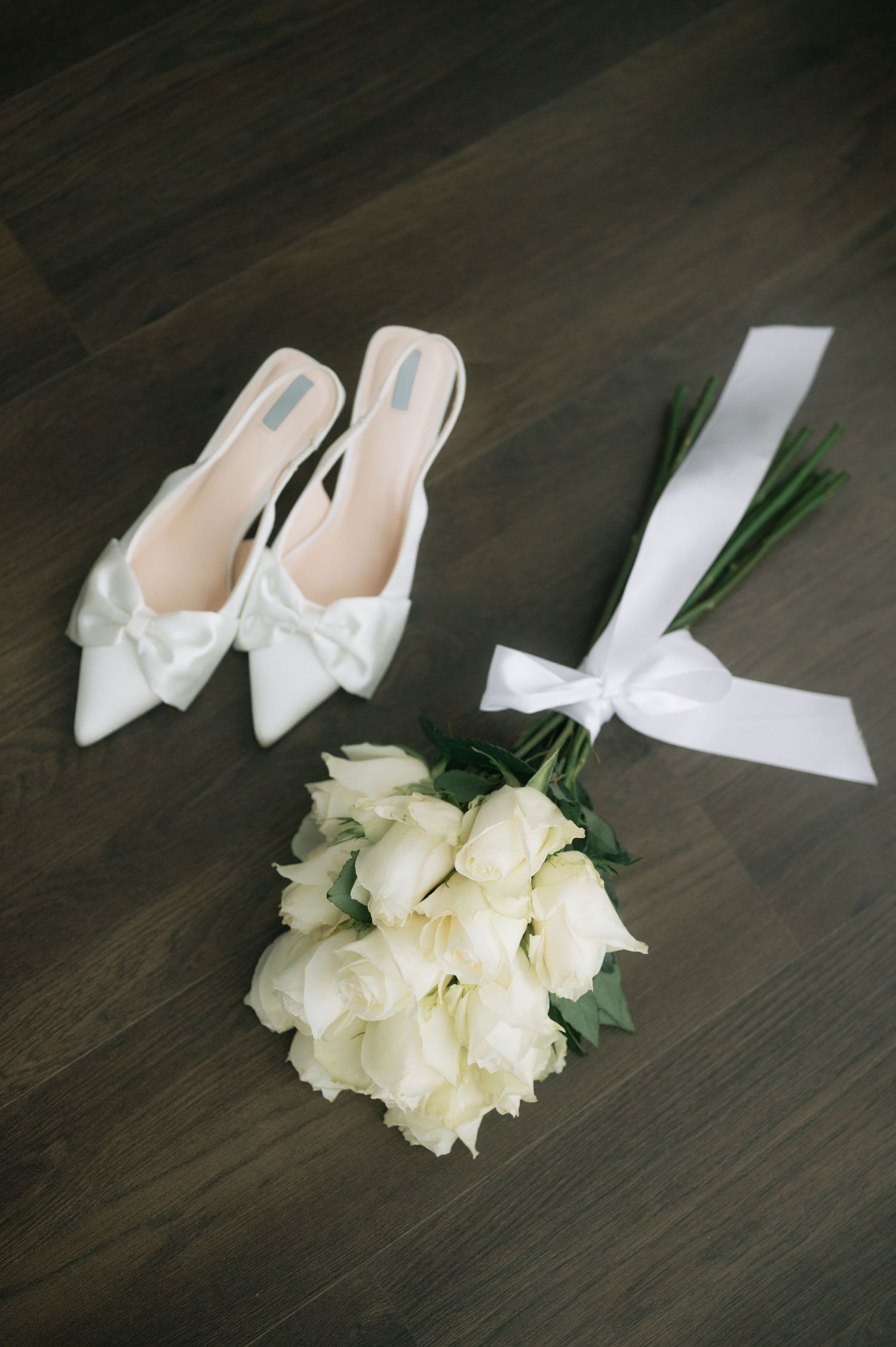White bridal shoes with bows displayed alongside a bouquet of white roses and a white ribbon on dark wooden flooring.