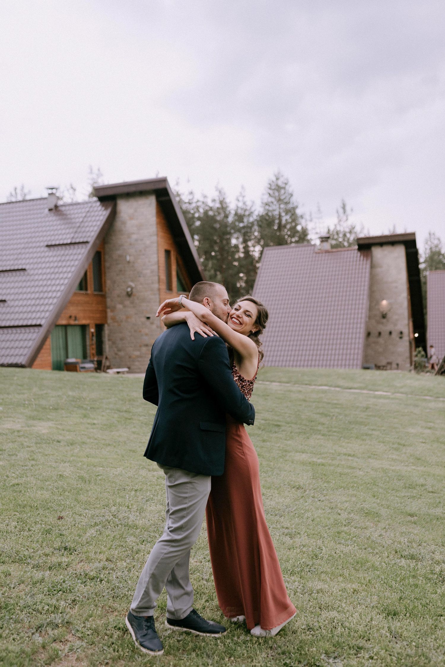Couple embraces on a grassy hill with A-frame cabins visible in the background.