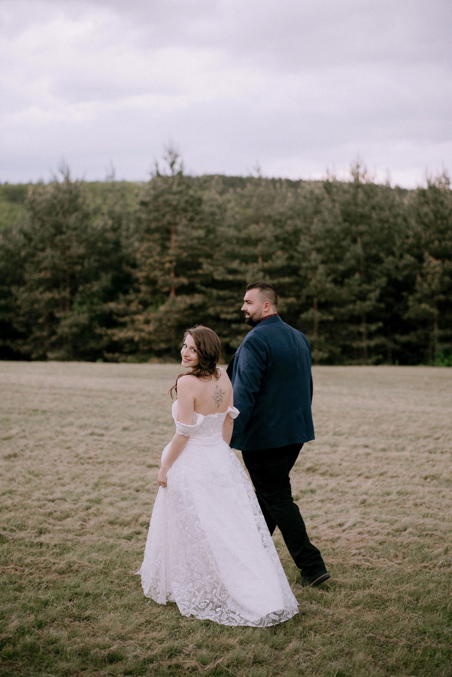 A couple in formal attire walks across a grassy hillside with pine forest in the background.