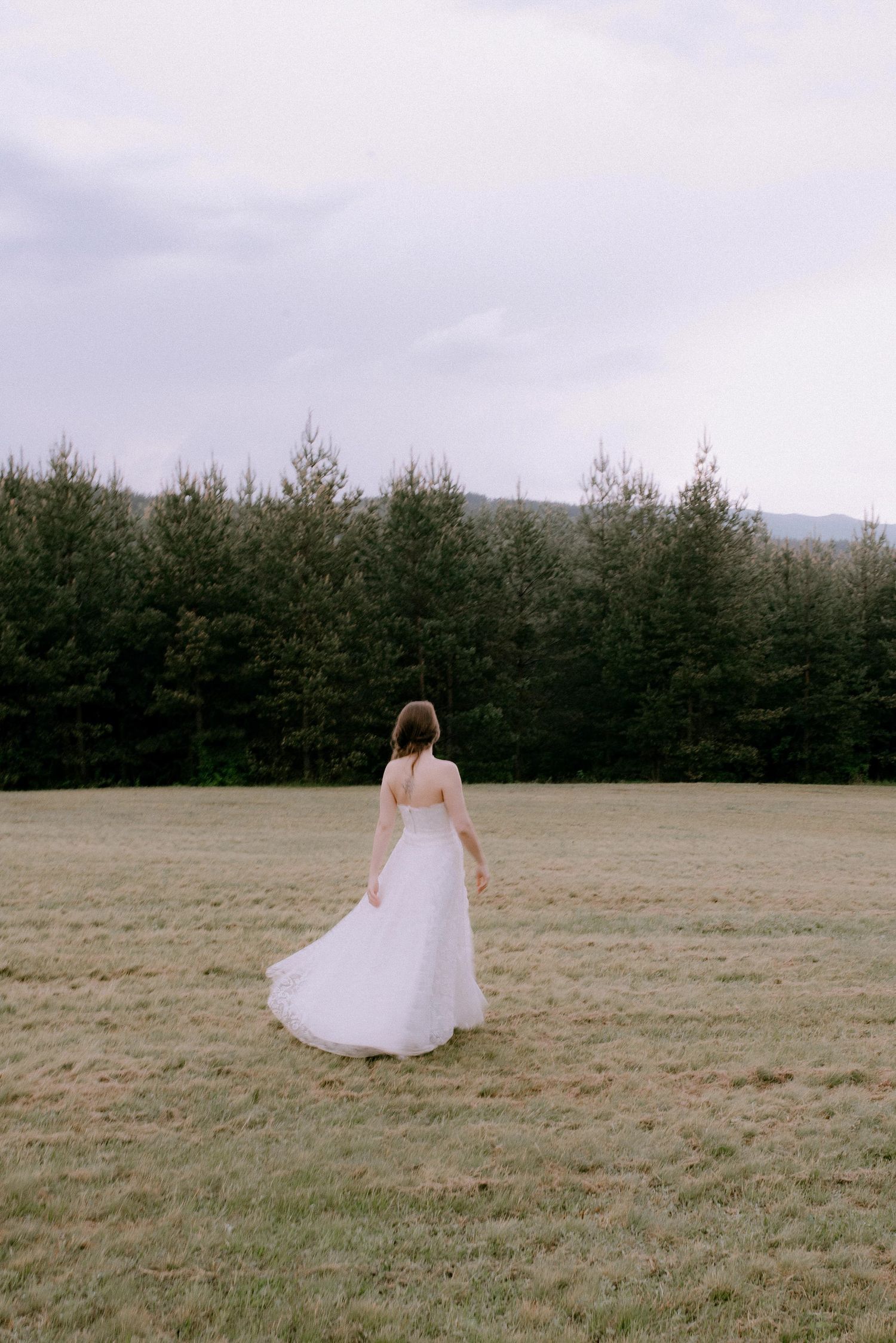 Person in a flowing white dress walking across a scenic meadow with forest backdrop.