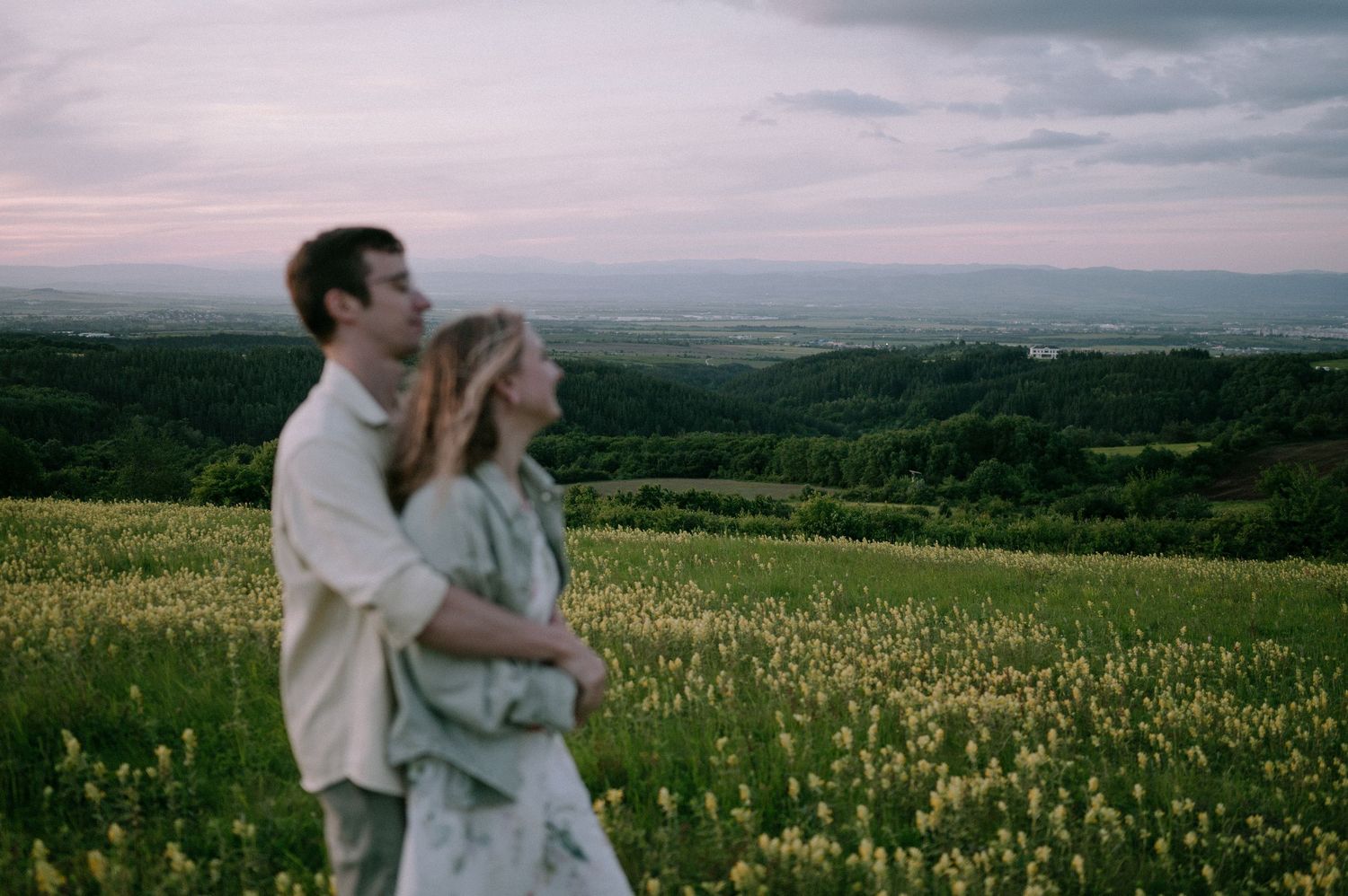 A couple embraces in a scenic wildflower meadow at sunset with rolling hills in the background.