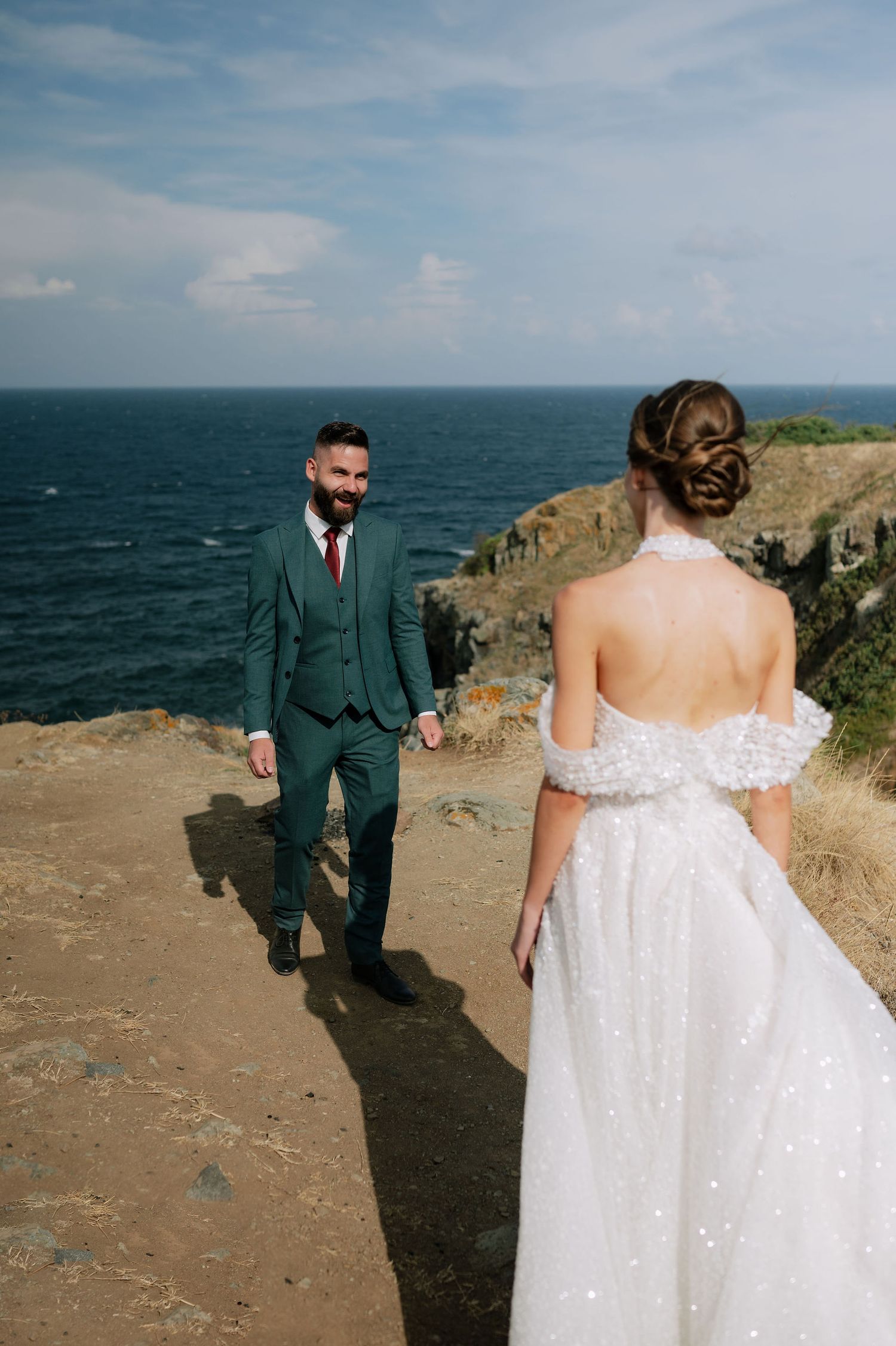 A couple in formal attire walking on a cliff path overlooking the ocean.