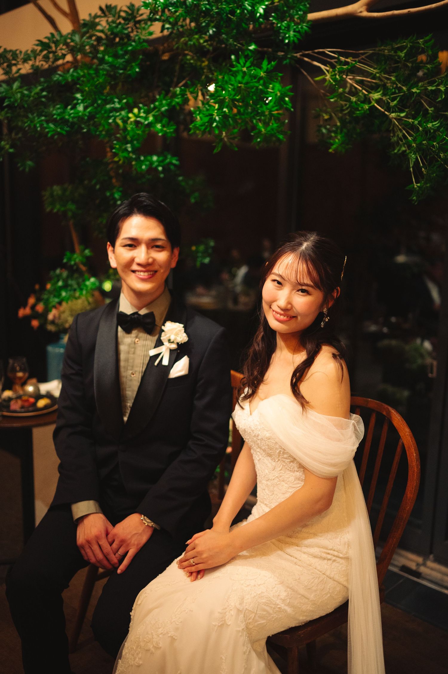A couple smiles during their wedding reception beneath glowing green branches.