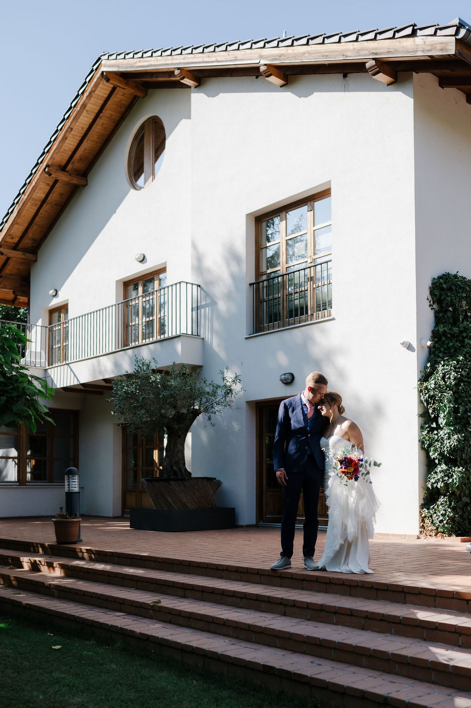 A wedding party stands outside a white Mediterranean-style building with terra cotta steps.