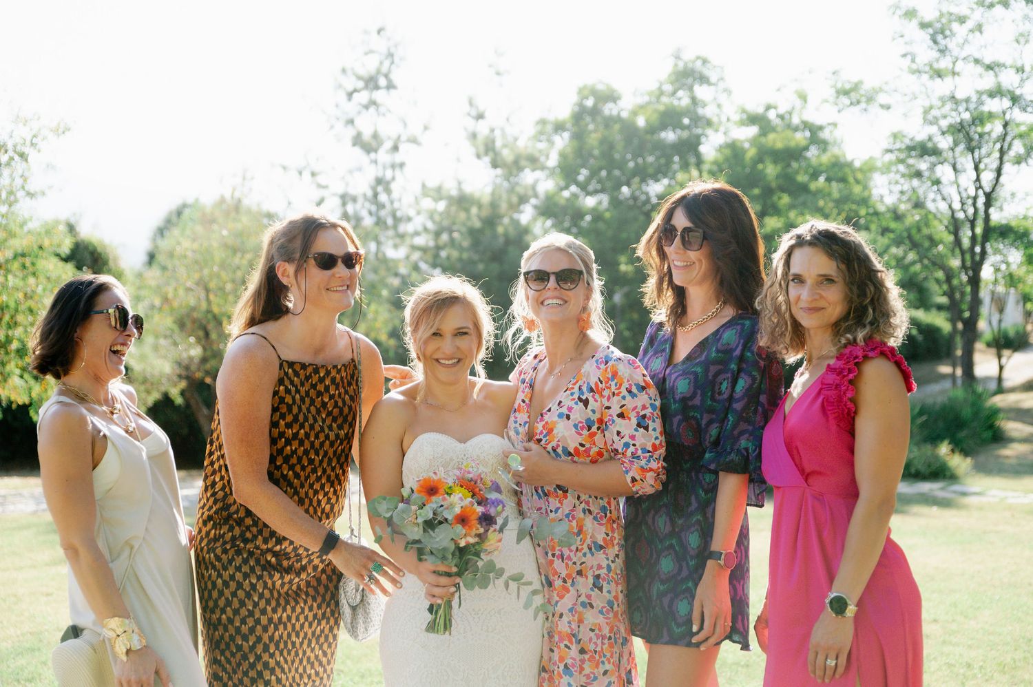 Wedding party members in bright summer dresses stand together outdoors.