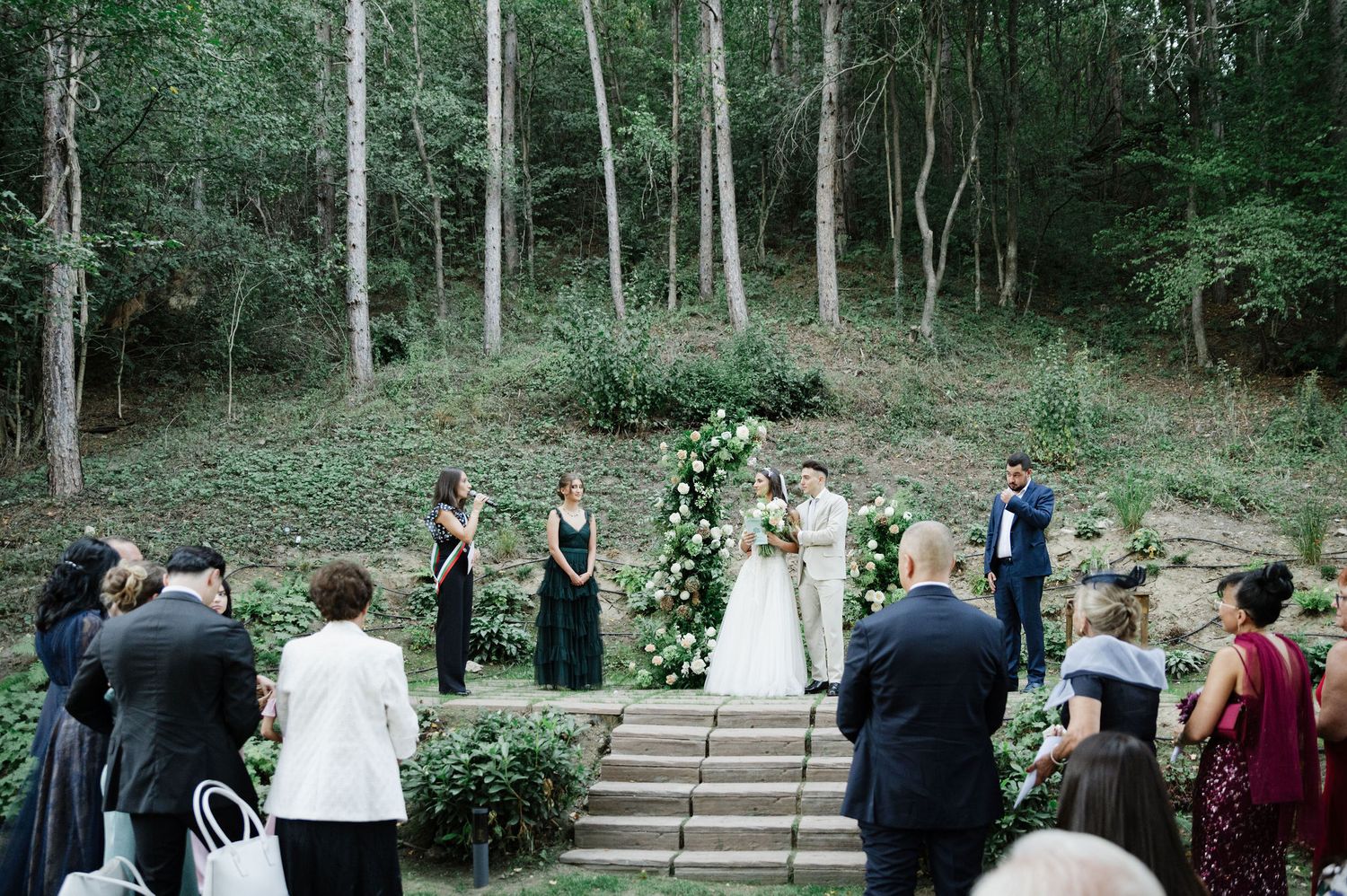 Wide view of forest wedding ceremony with guests gathered around stone steps and floral arrangements.