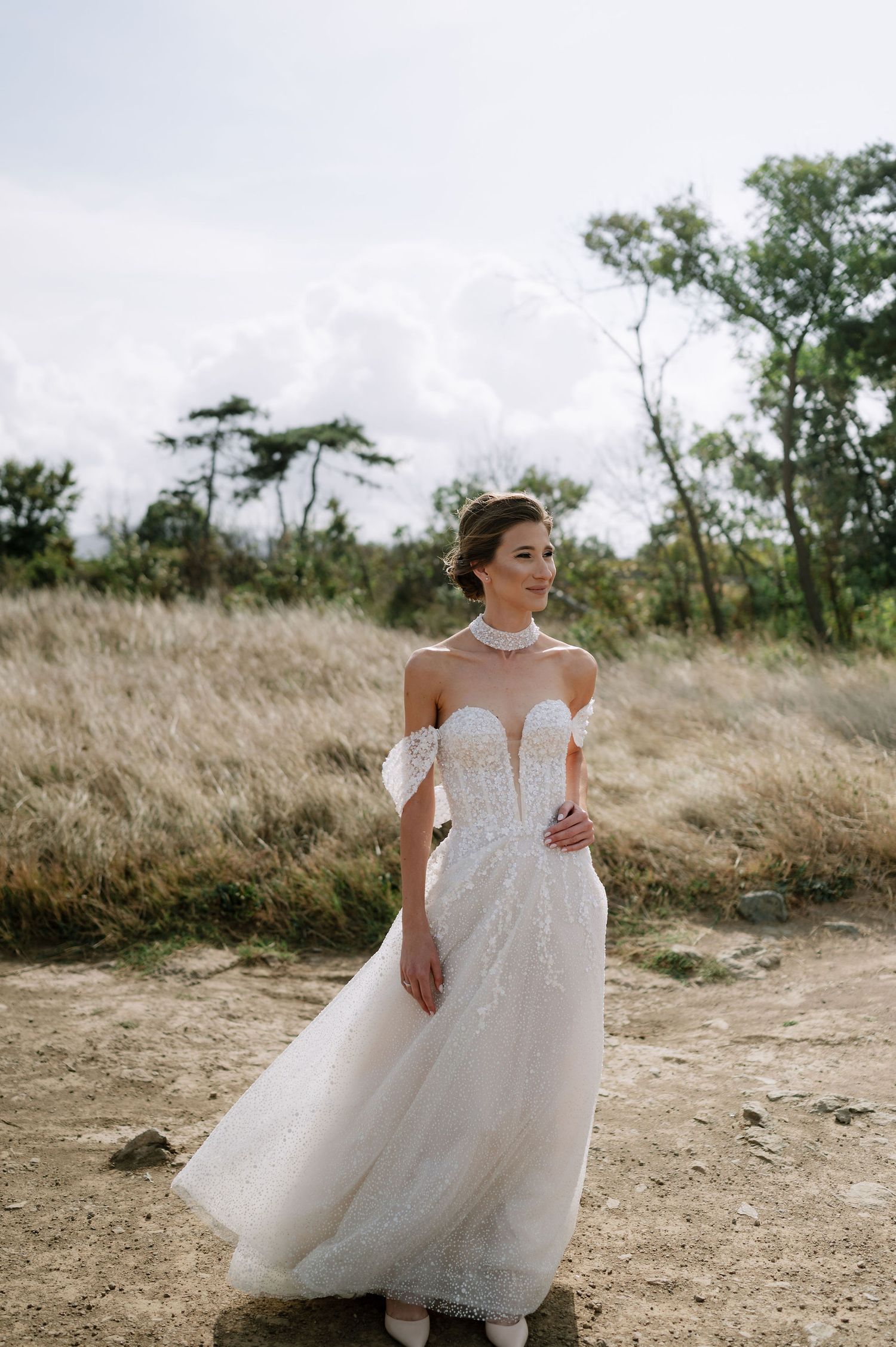 Bride in flowing white gown twirls on natural path surrounded by tall grass and trees.