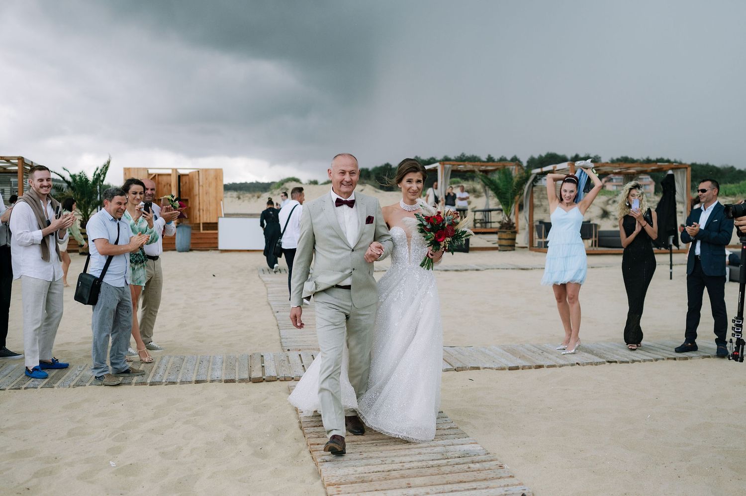 Wedding guests gather on a beach boardwalk under cloudy skies during an outdoor ceremony.
