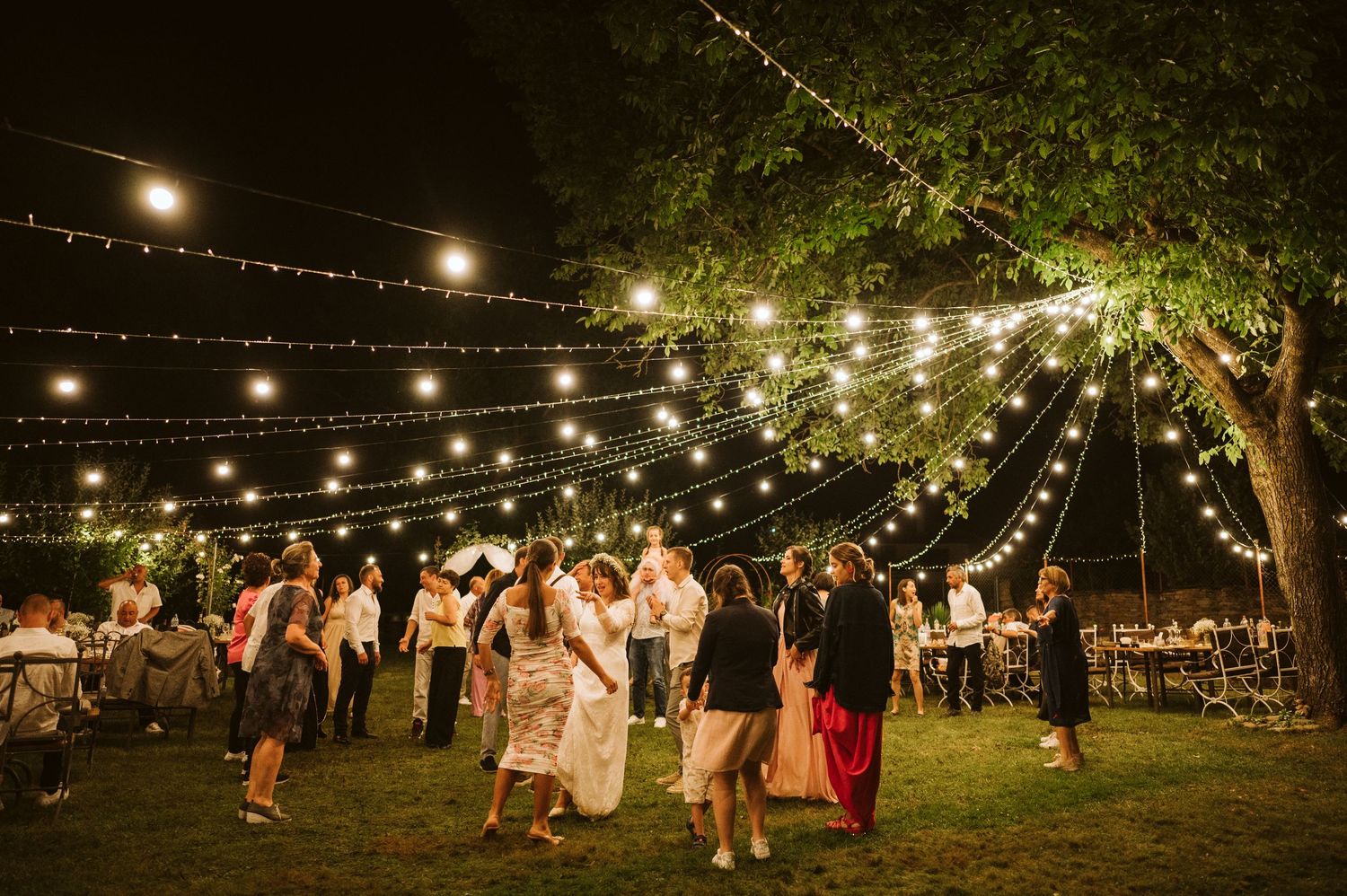 Wedding guests dance under romantic string lights strung between trees at an outdoor evening reception.