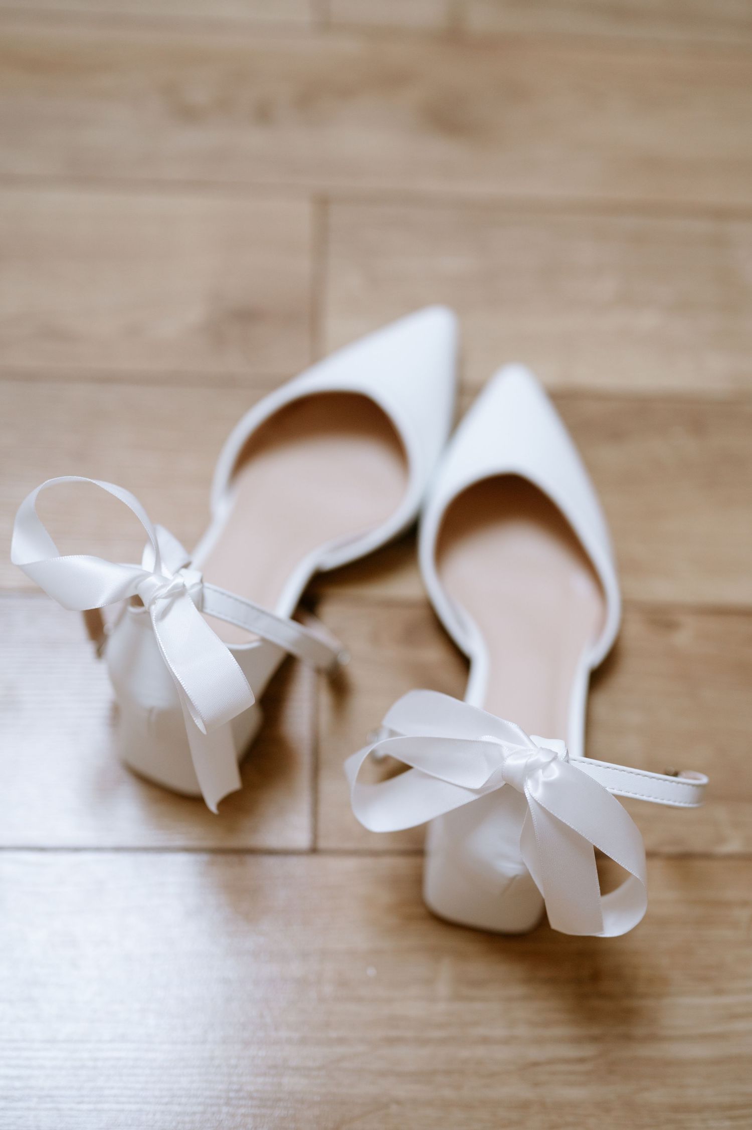White pointed-toe wedding flats with delicate bow details on wooden flooring.