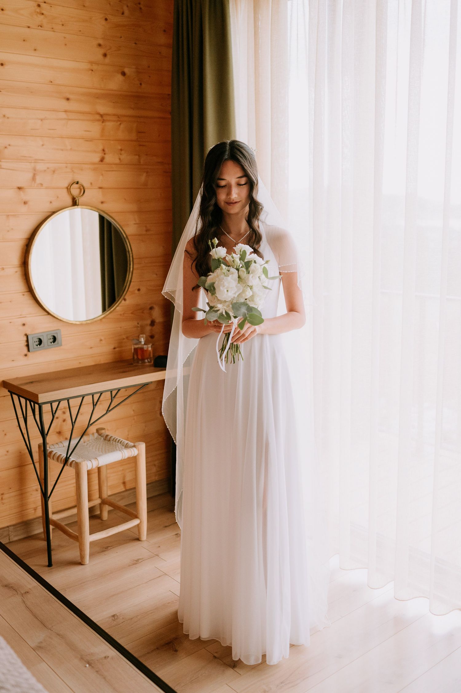 Elegant bridal portrait in white dress against wood paneled walls and sheer curtains.