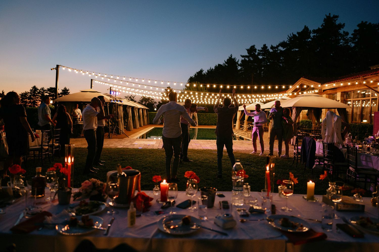 String lights illuminate an outdoor evening reception space with tables and chairs set up on a lawn.