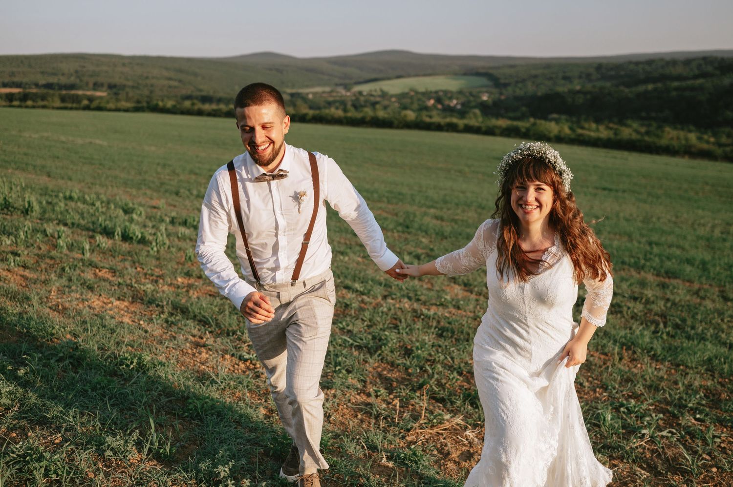 A couple dressed in white runs joyfully through a rolling green meadow with mountains in the background.
