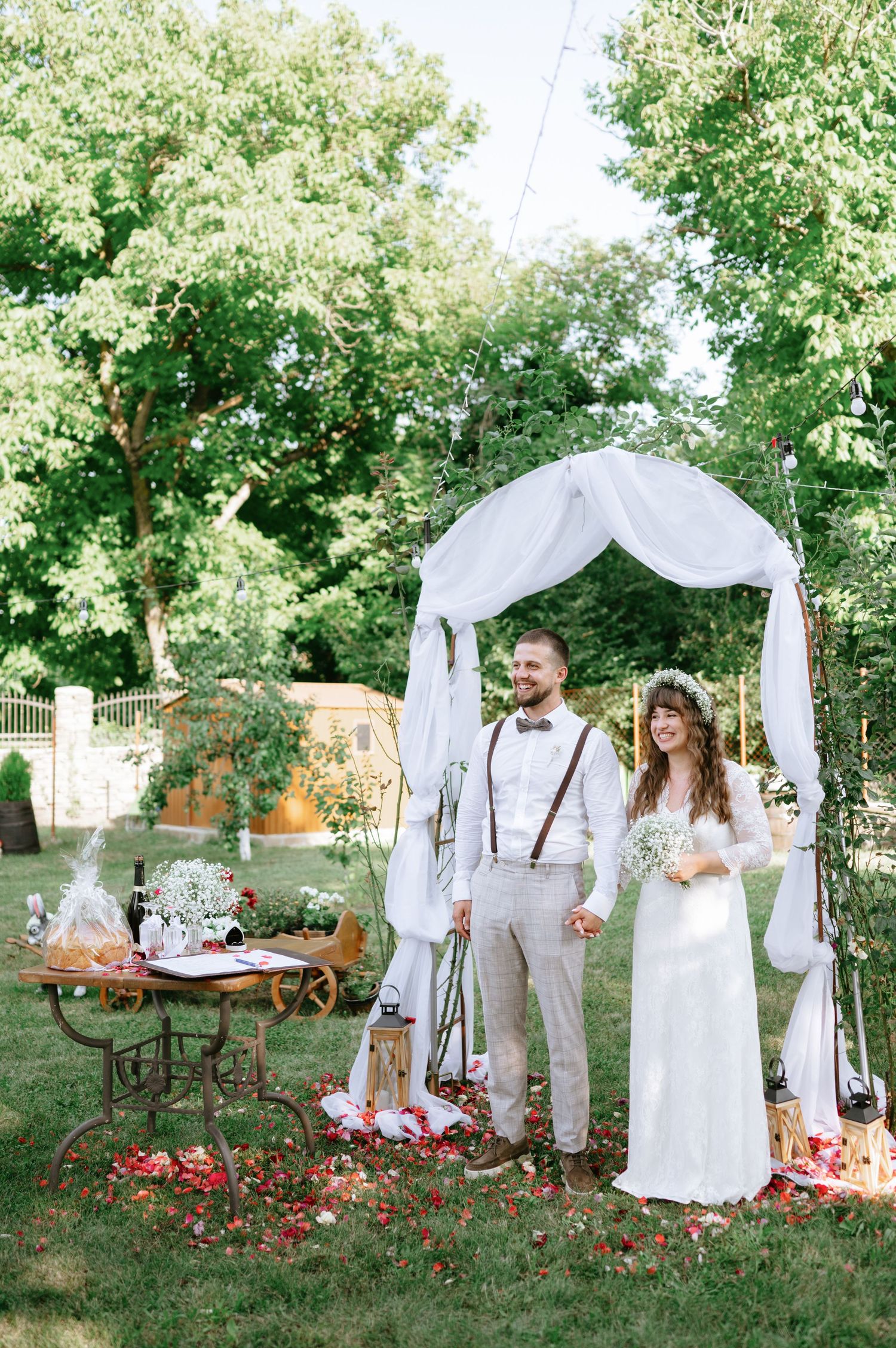 Outdoor wedding venue with white fabric arch, vintage furniture and scattered rose petals in natural setting.