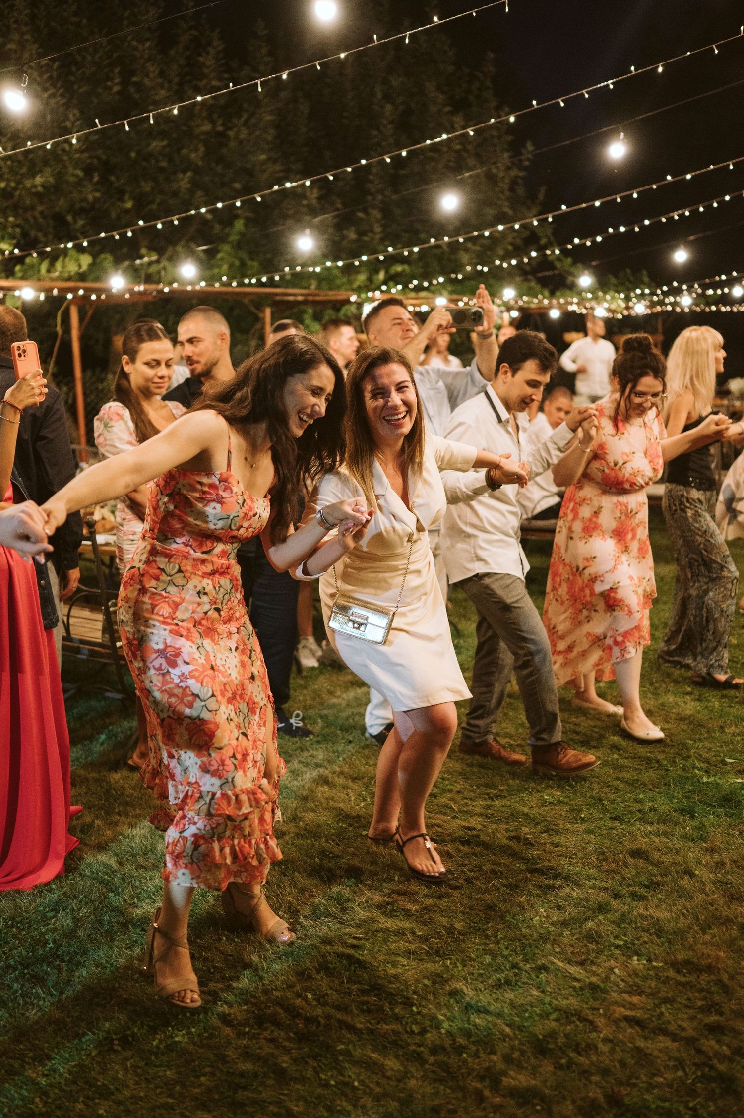 Wedding guests dancing energetically on grass under string lights at outdoor reception.