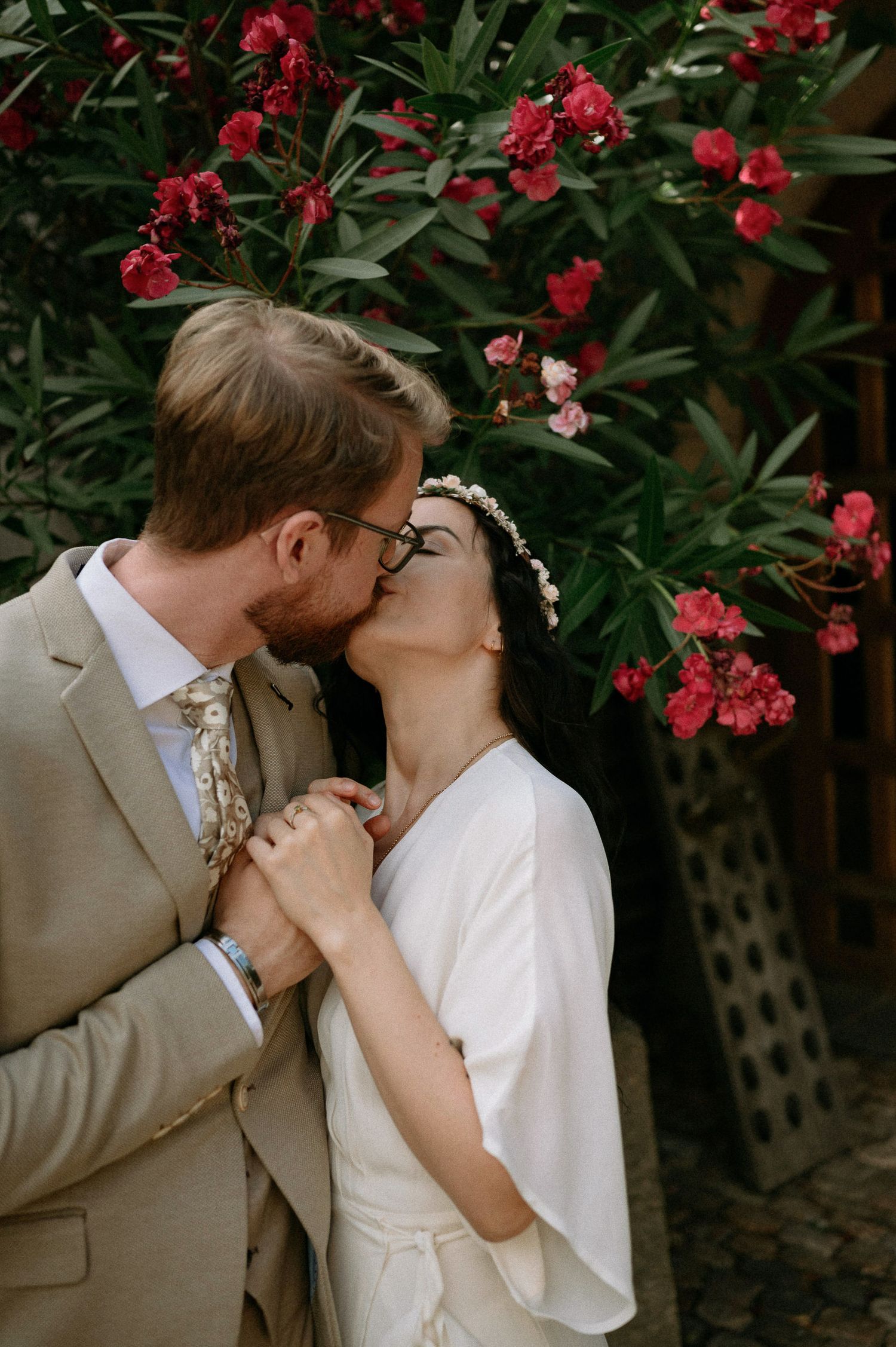 Intimate wedding kiss captured in front of vibrant red flowering bushes.