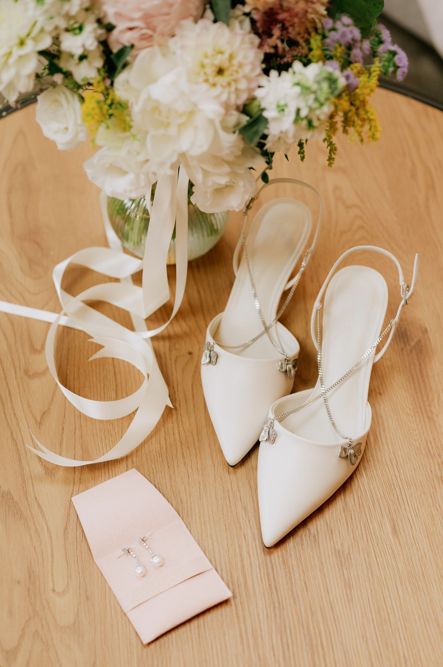 Elegant white wedding shoes and floral arrangement on light wood table.