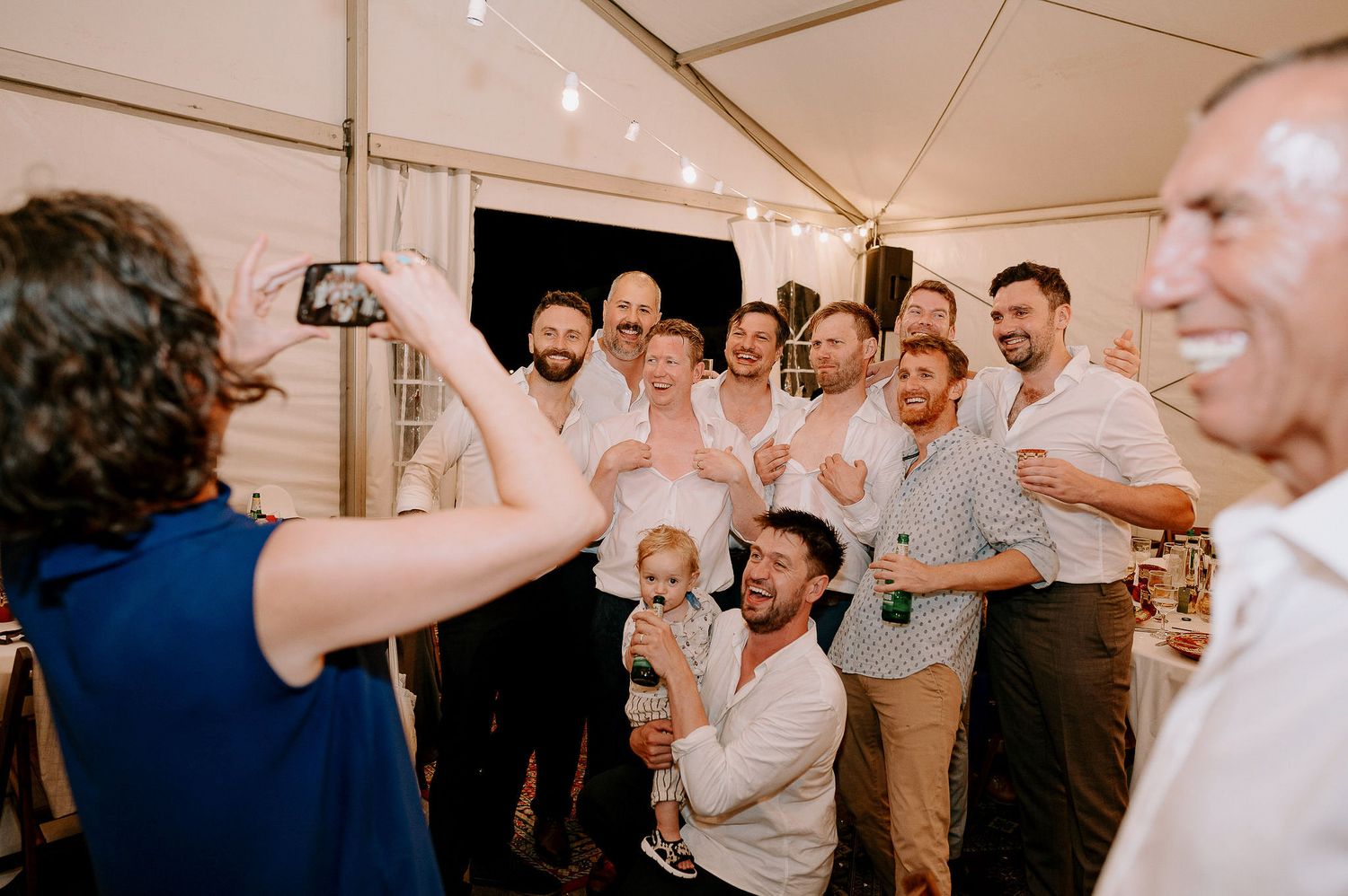 Energetic crowd dancing and celebrating at a wedding reception under white tent lighting.