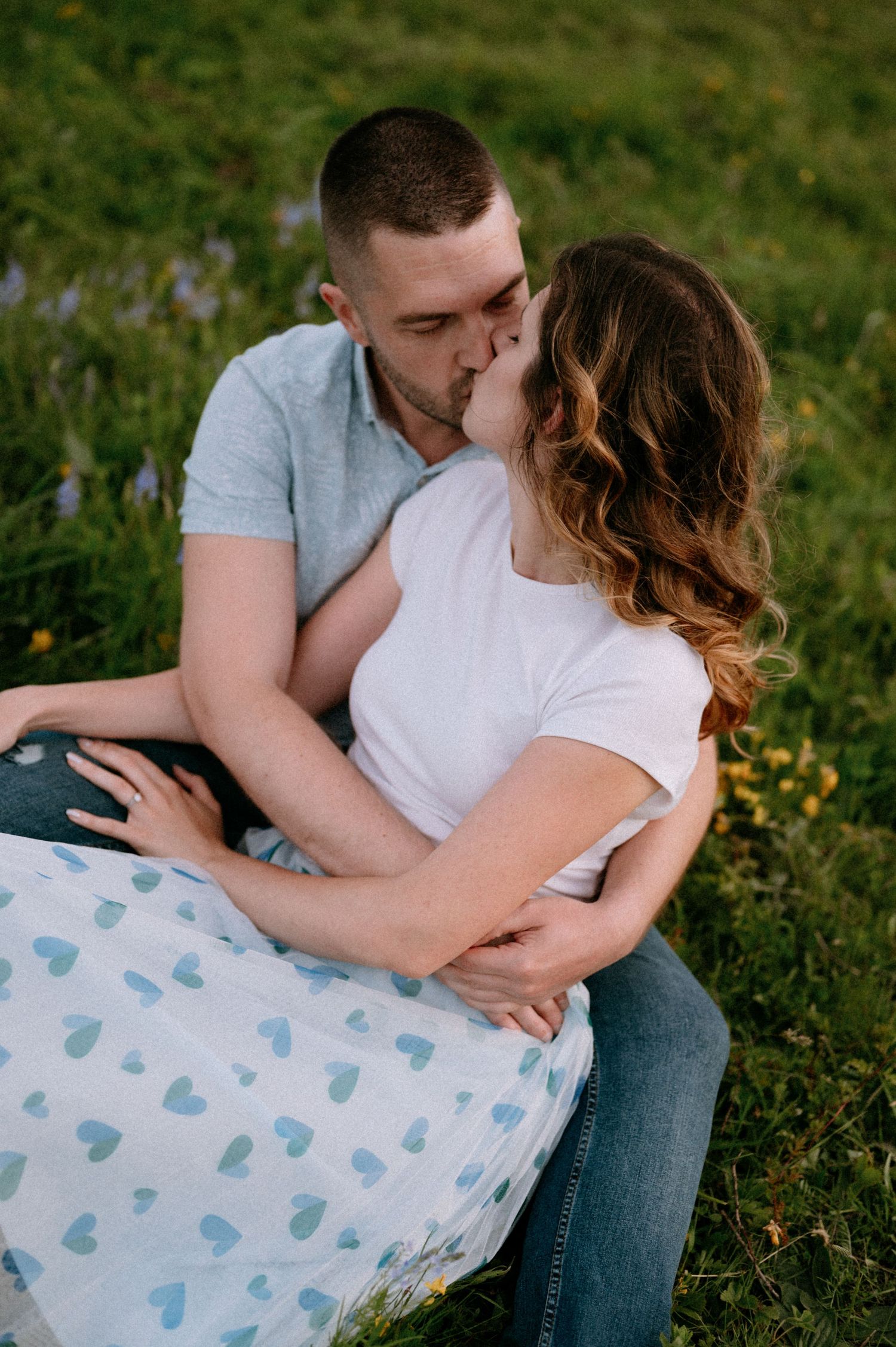 Couple cuddling on blanket with heart pattern in mountain meadow surrounded by flowers.