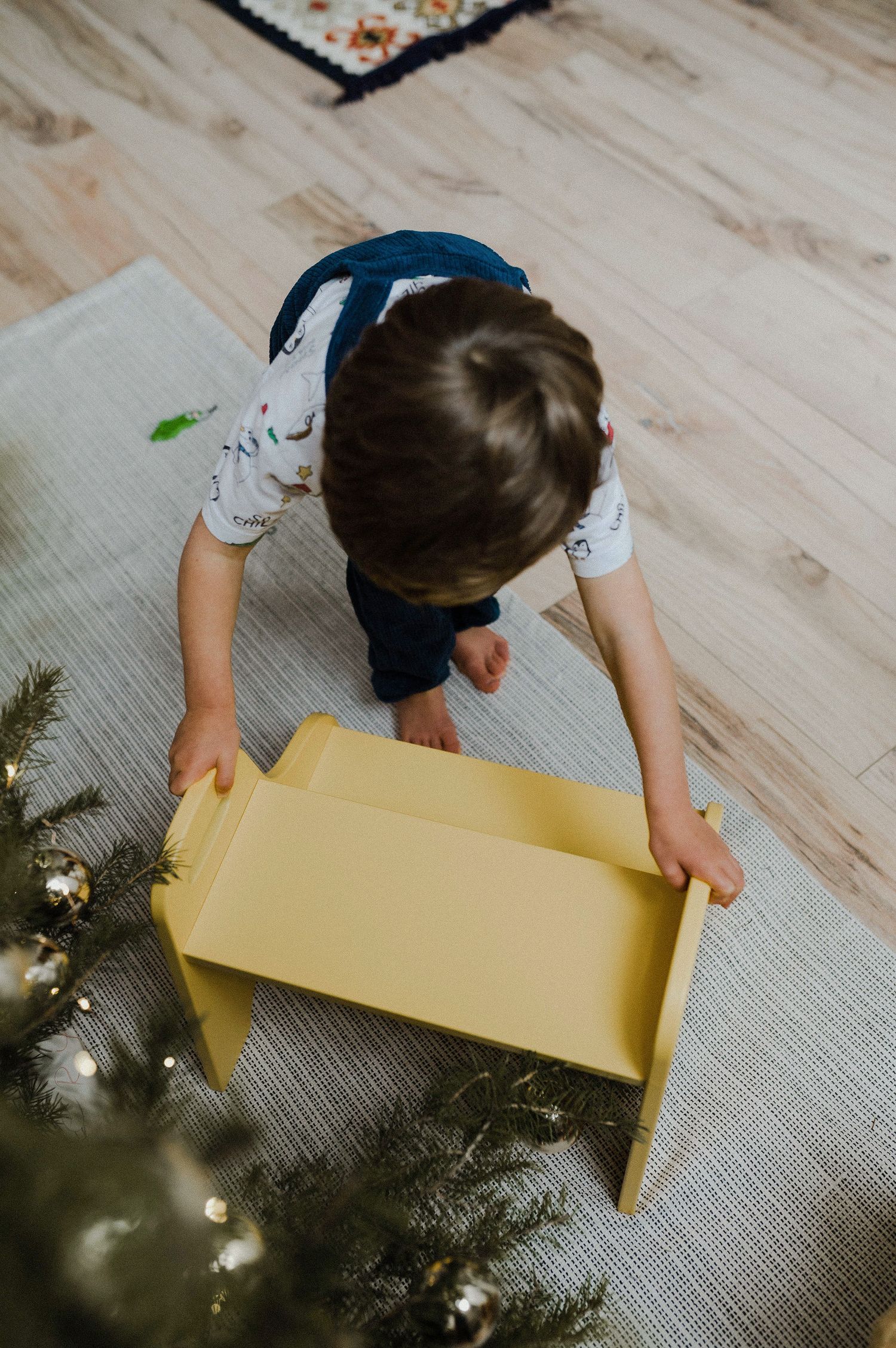 Overhead view of someone reaching into a yellow box next to a decorated Christmas tree on a light wooden floor.
