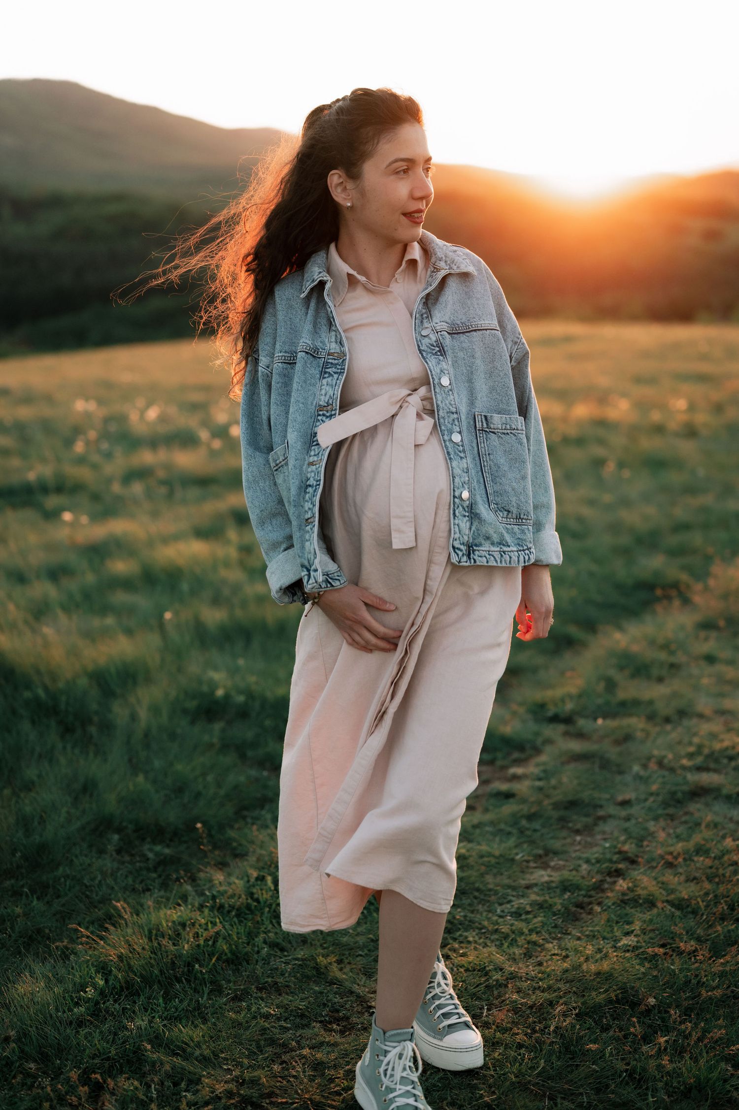 A person wearing a beige dress and jean jacket poses playfully in a sunlit meadow during golden hour.
