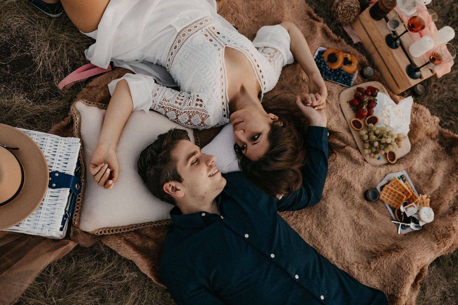 Overhead view of couple lying on picnic blanket with snacks and drinks.