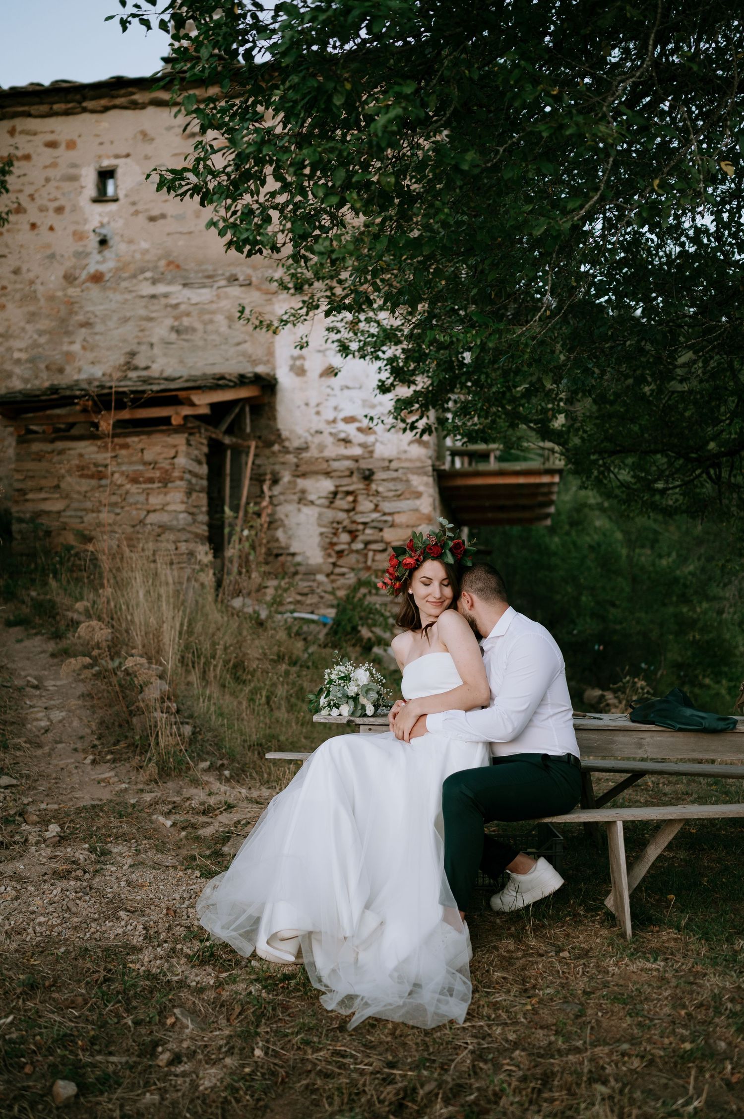 A romantic wedding moment captured on a rustic wooden bench near an old stone wall.