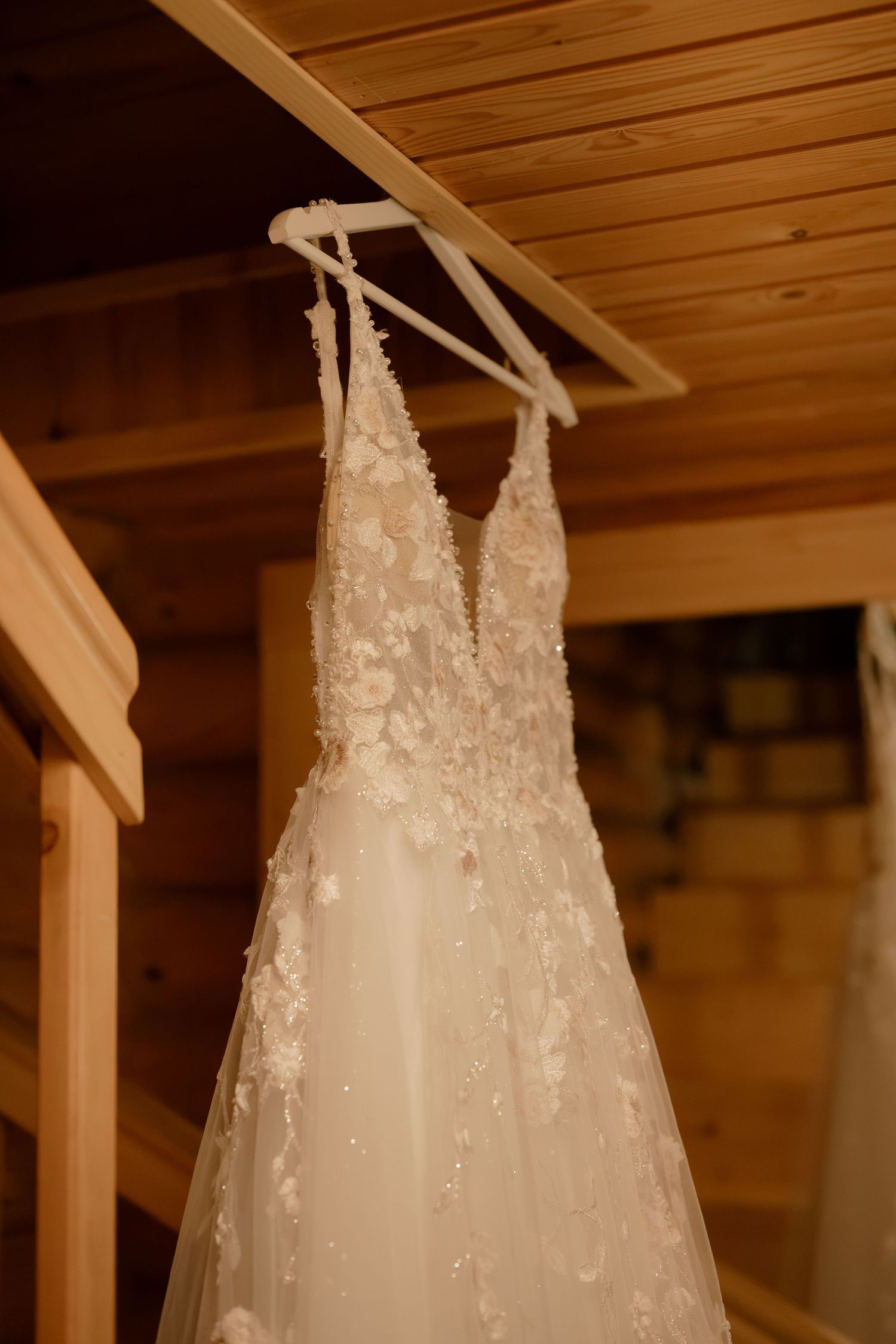 Elegant lace wedding dress with floral appliques hanging on a white hanger in a warm wooden cabin interior.
