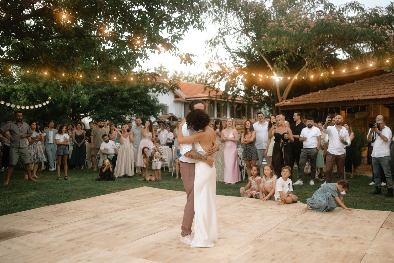 Couple shares first dance at outdoor wedding reception with string lights and guests watching.
