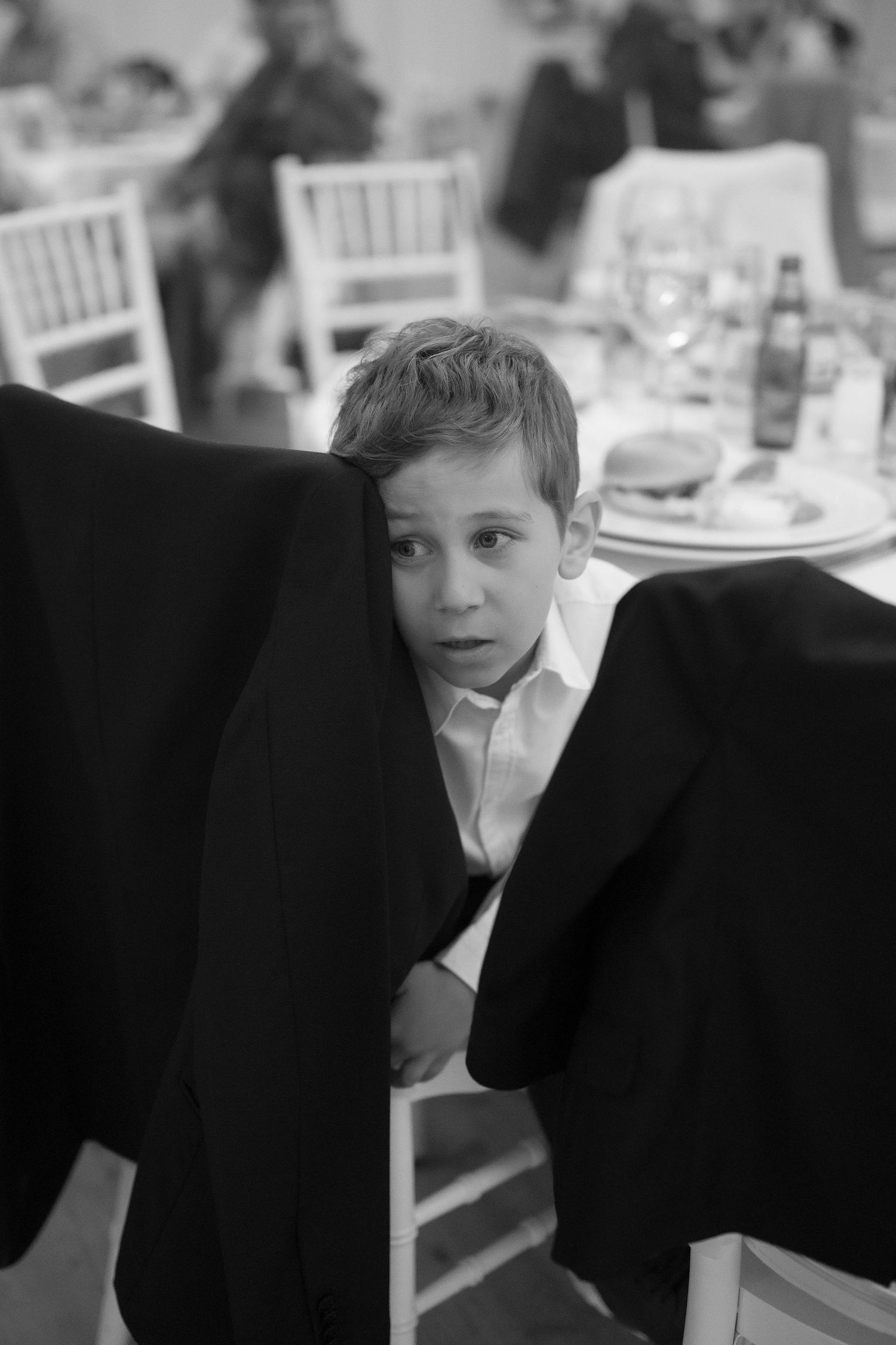 Black and white photo of a young boy in a dress shirt peeking between two dark suit jackets at a formal event.