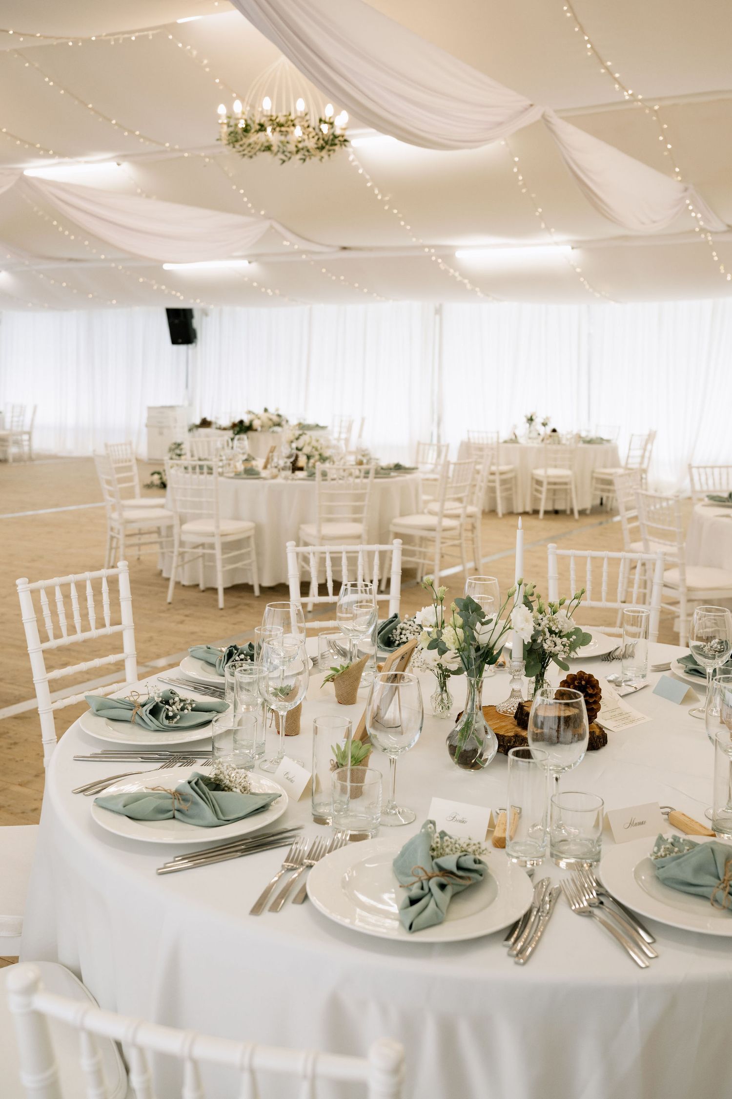 Elegant wedding reception tent with white Chiavari chairs, sage green napkins, floral centerpieces, and fairy lights draped from ceiling.