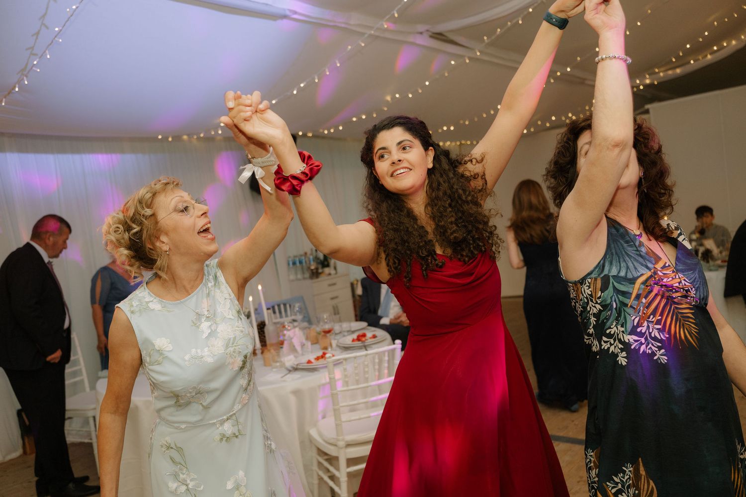 Three women dancing joyfully at a wedding reception, raising hands together in elegant dresses under warm string lights.