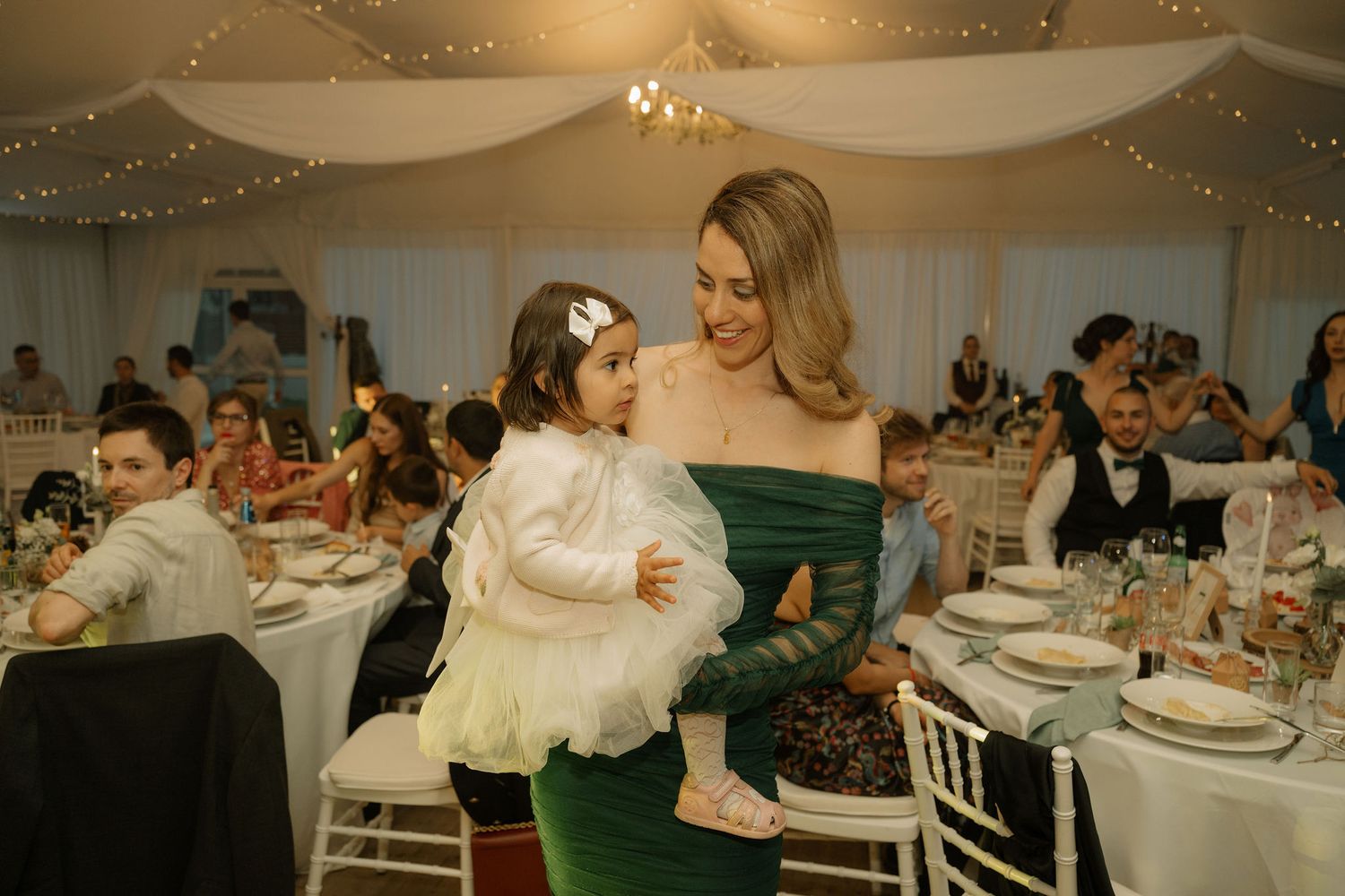 Woman in green dress holds young girl in white dress at elegant wedding reception venue.