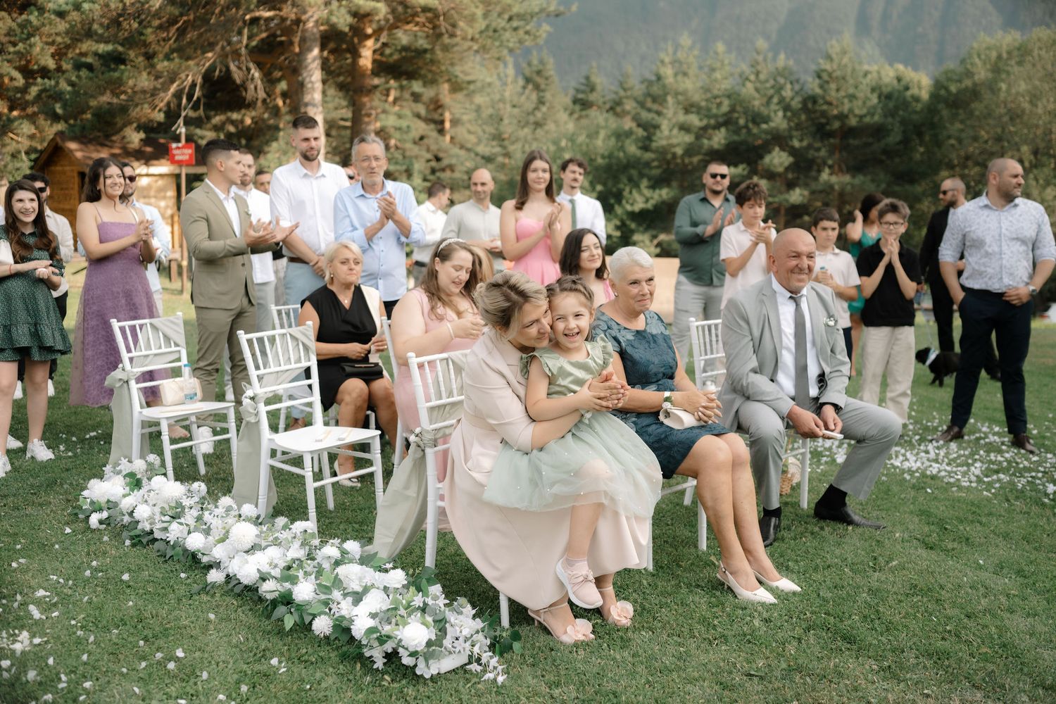 Wedding guests seated on white chairs in an outdoor garden ceremony with floral arrangements lining the aisle.