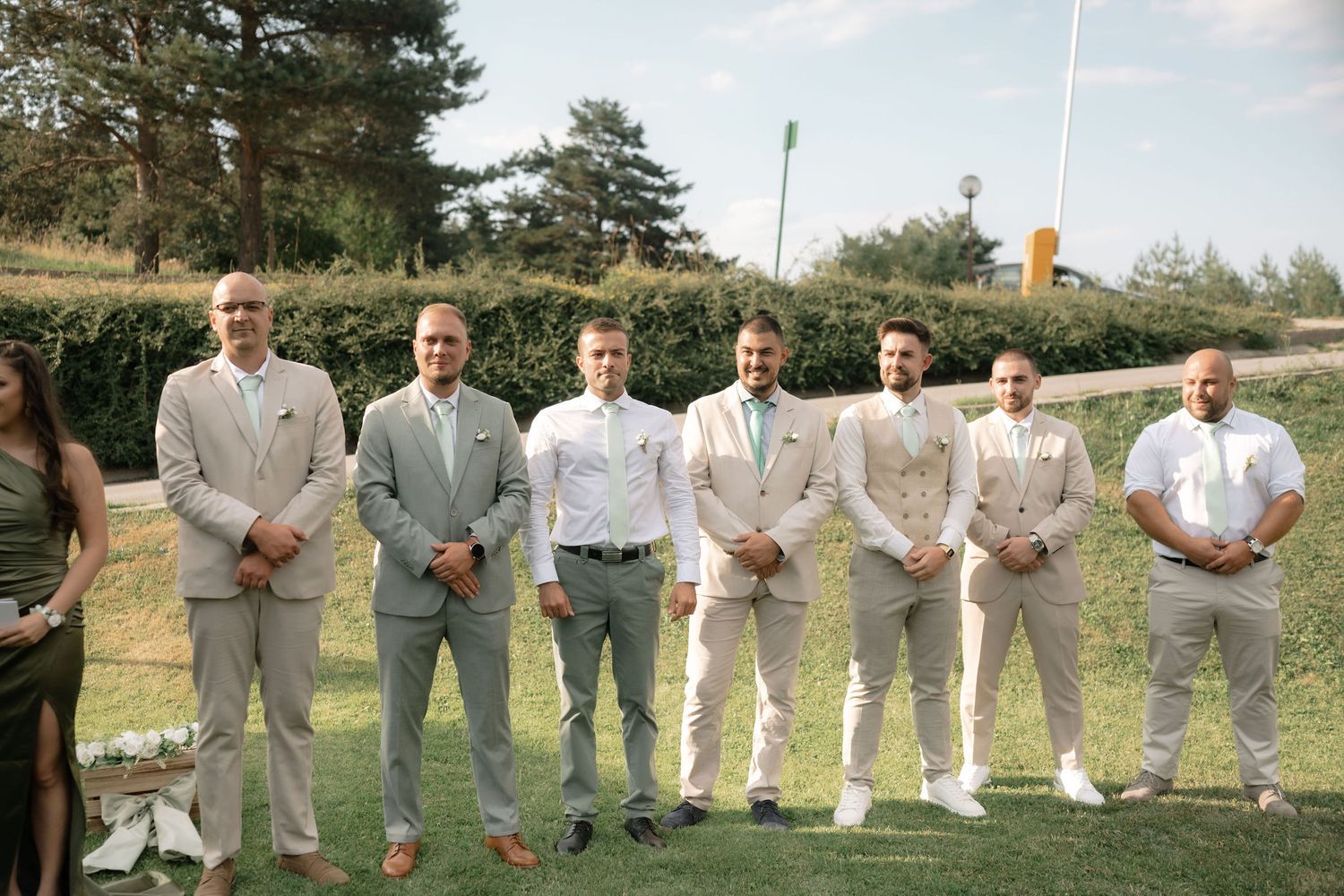 Wedding party in light gray and white attire poses together in a row on grassy outdoor hillside.