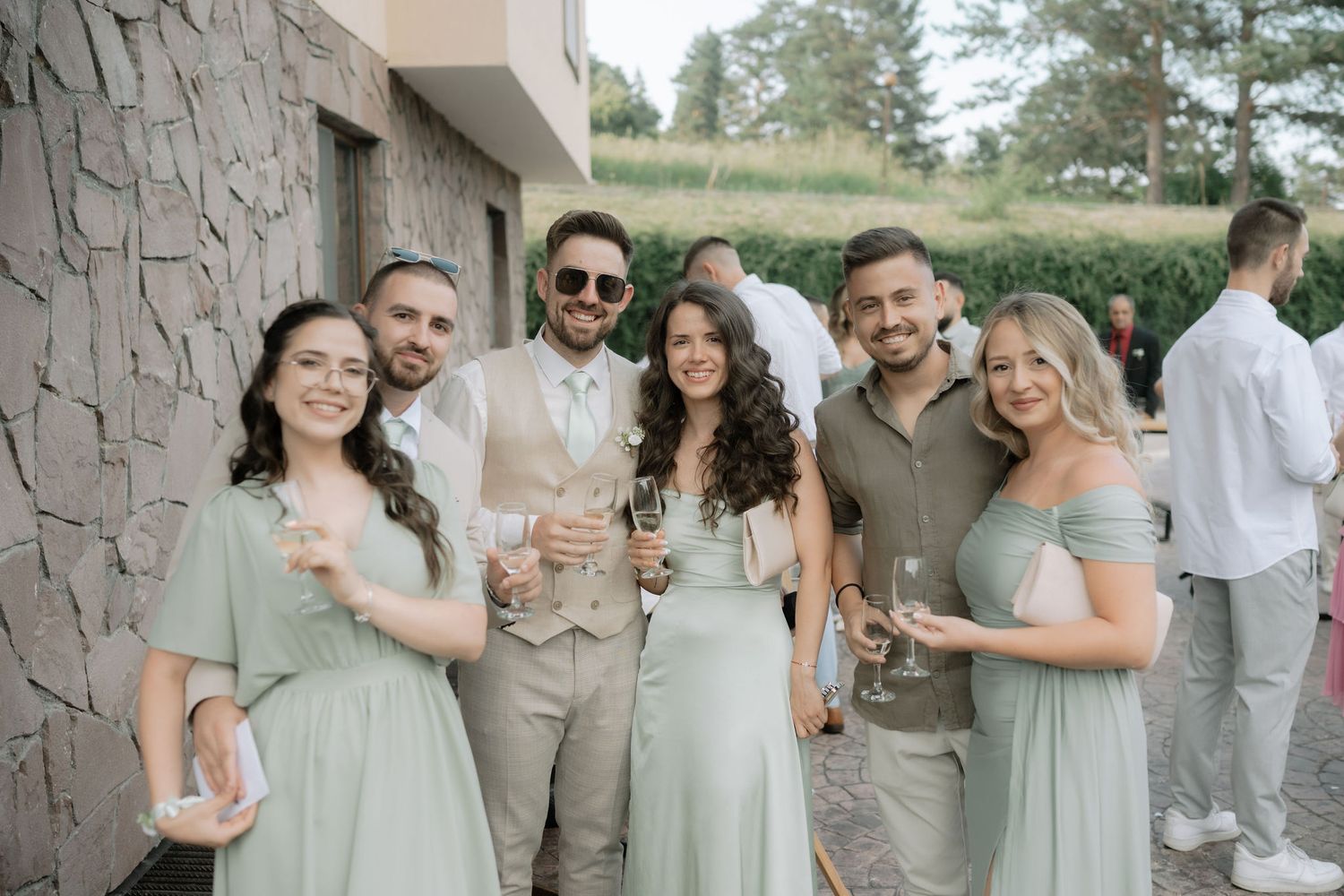 Wedding party in coordinated sage green and gray outfits poses against stone wall backdrop at outdoor venue.