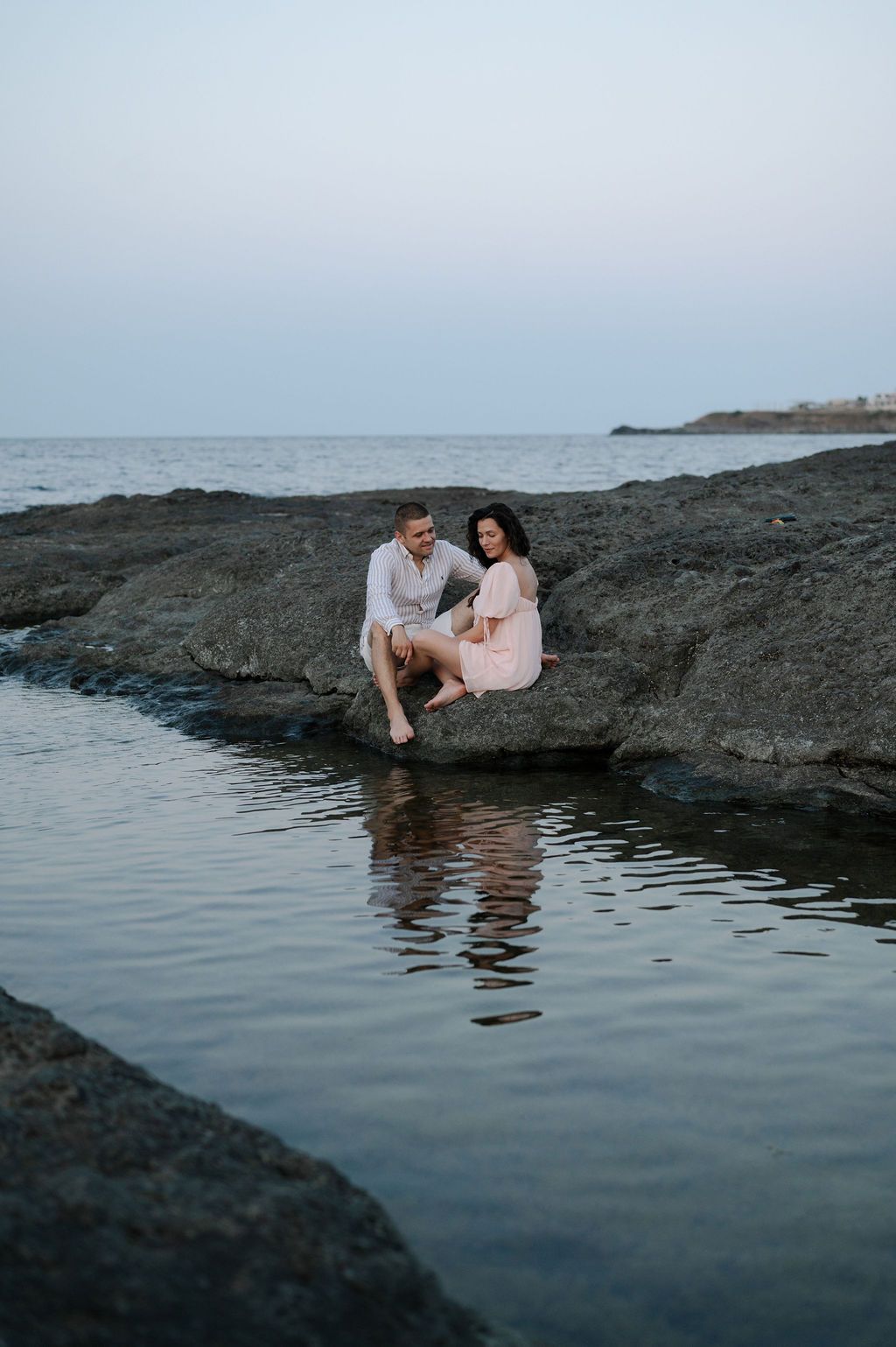 A couple sits together on coastal rocks at dusk, their reflection visible in the calm water below.