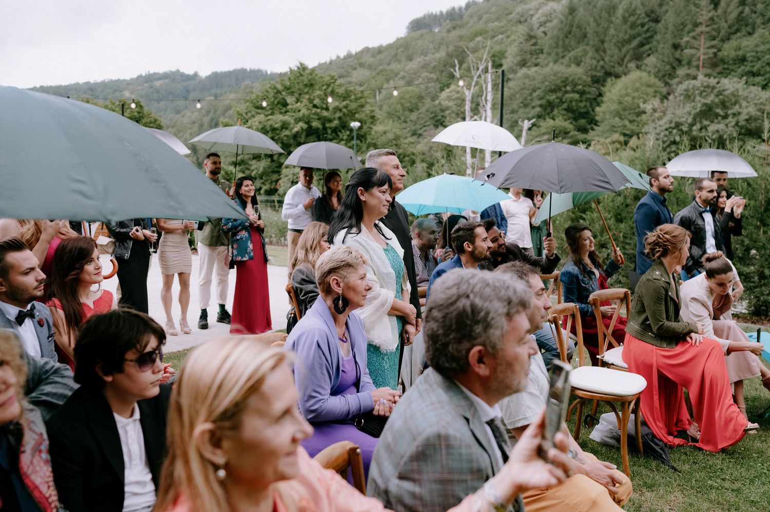 A crowd gathers outdoors under umbrellas and tents for a social event in a mountainous rural setting.