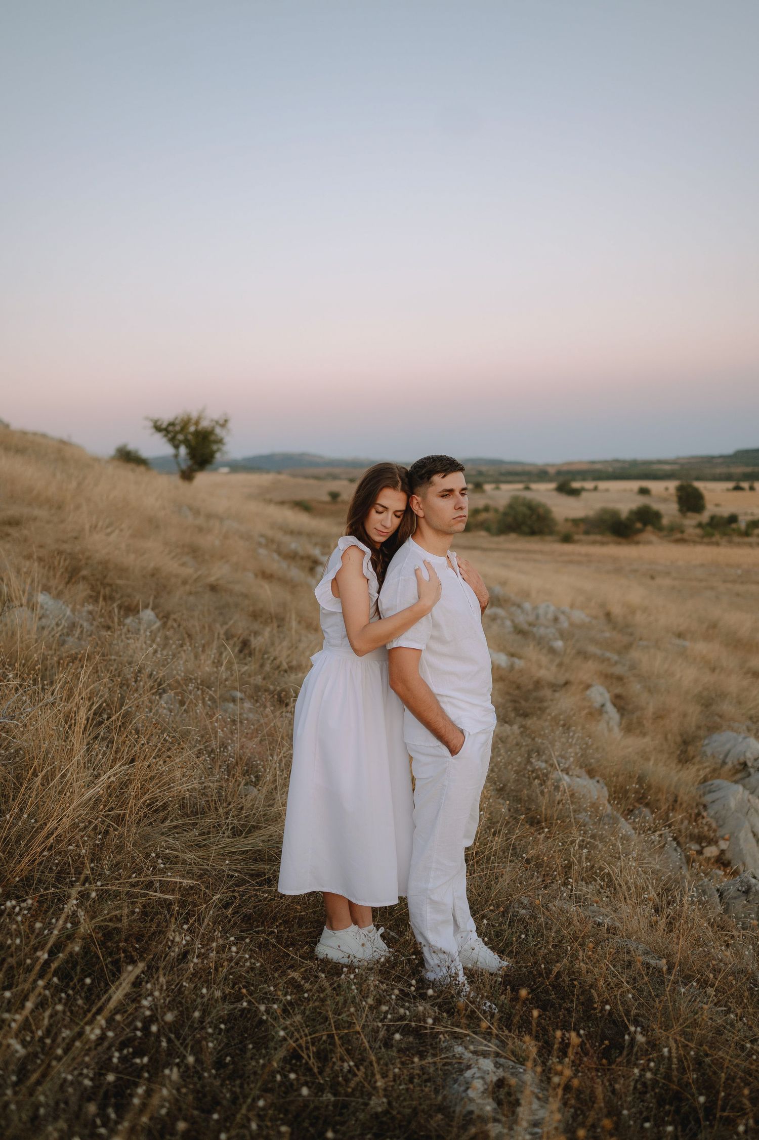 A couple in white outfits embraces on a hillside during golden hour with dry grass and mountains in the background.