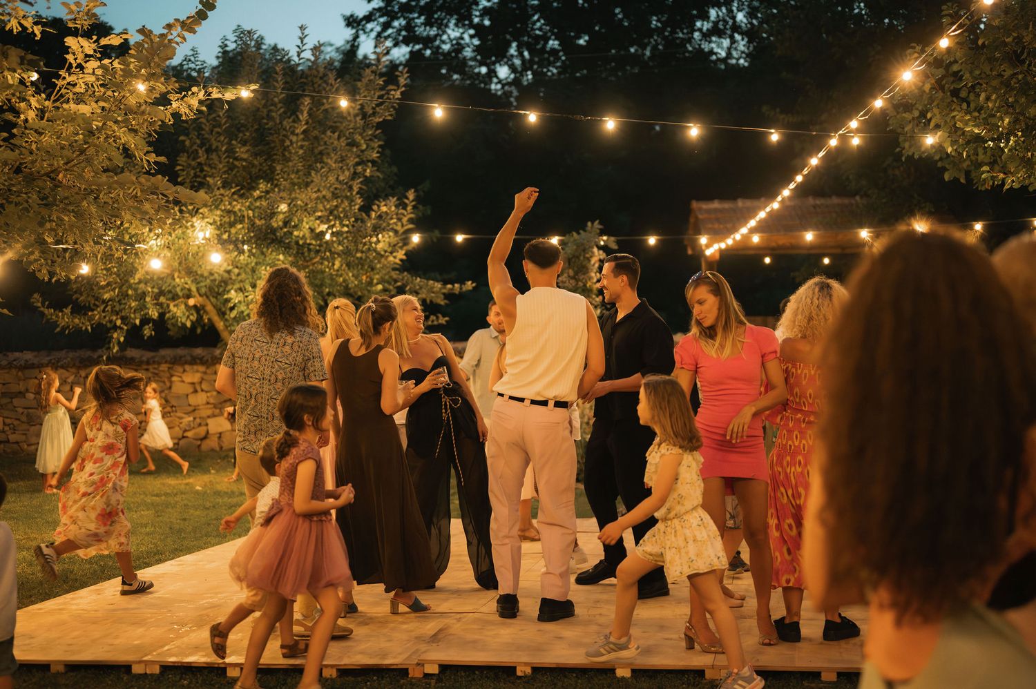 String lights illuminate an outdoor evening party as people dance and socialize on a warm summer night.